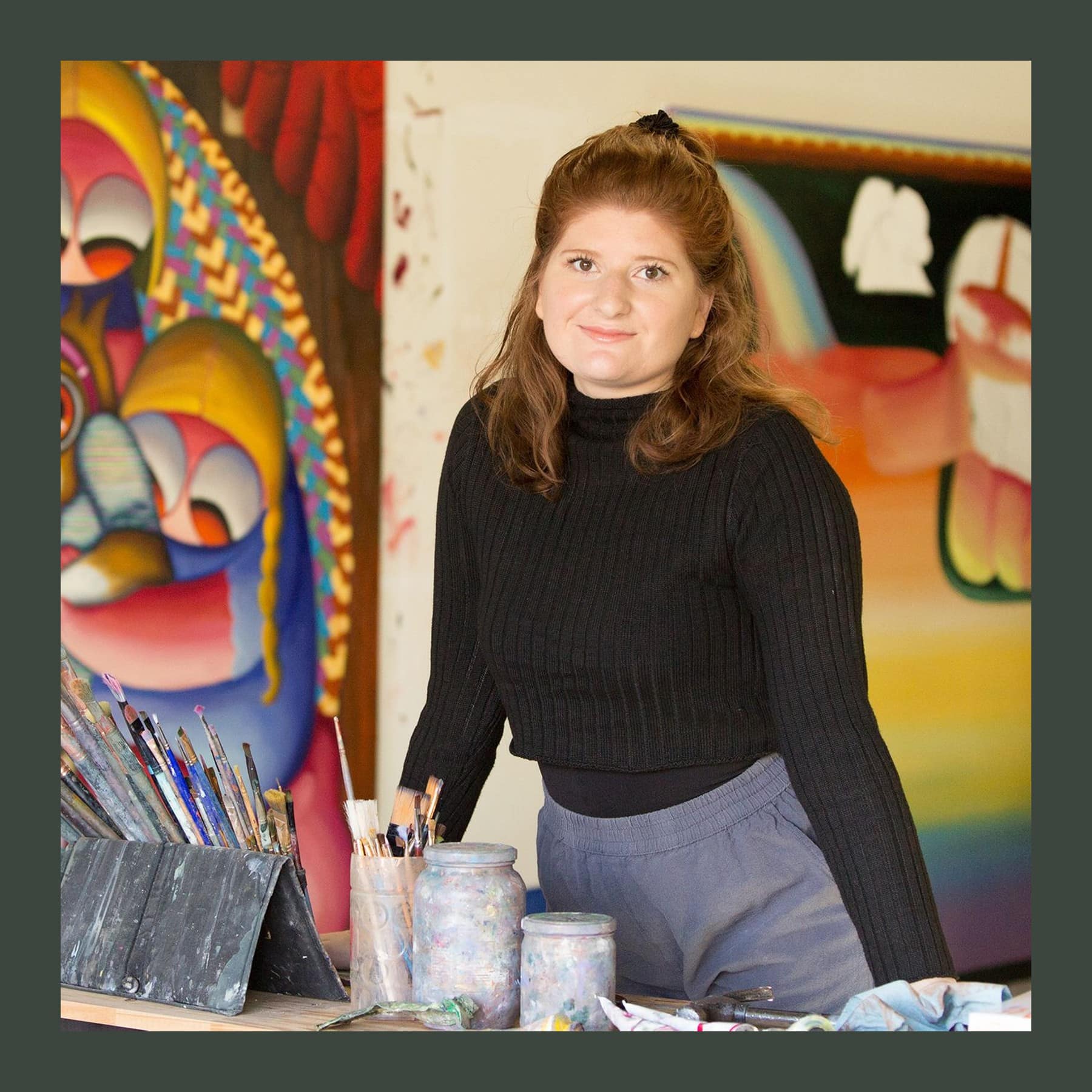 Natalie Wadlington in a black jumper at her studio table with brushes and jars, between two of her large, brightly coloured paintings