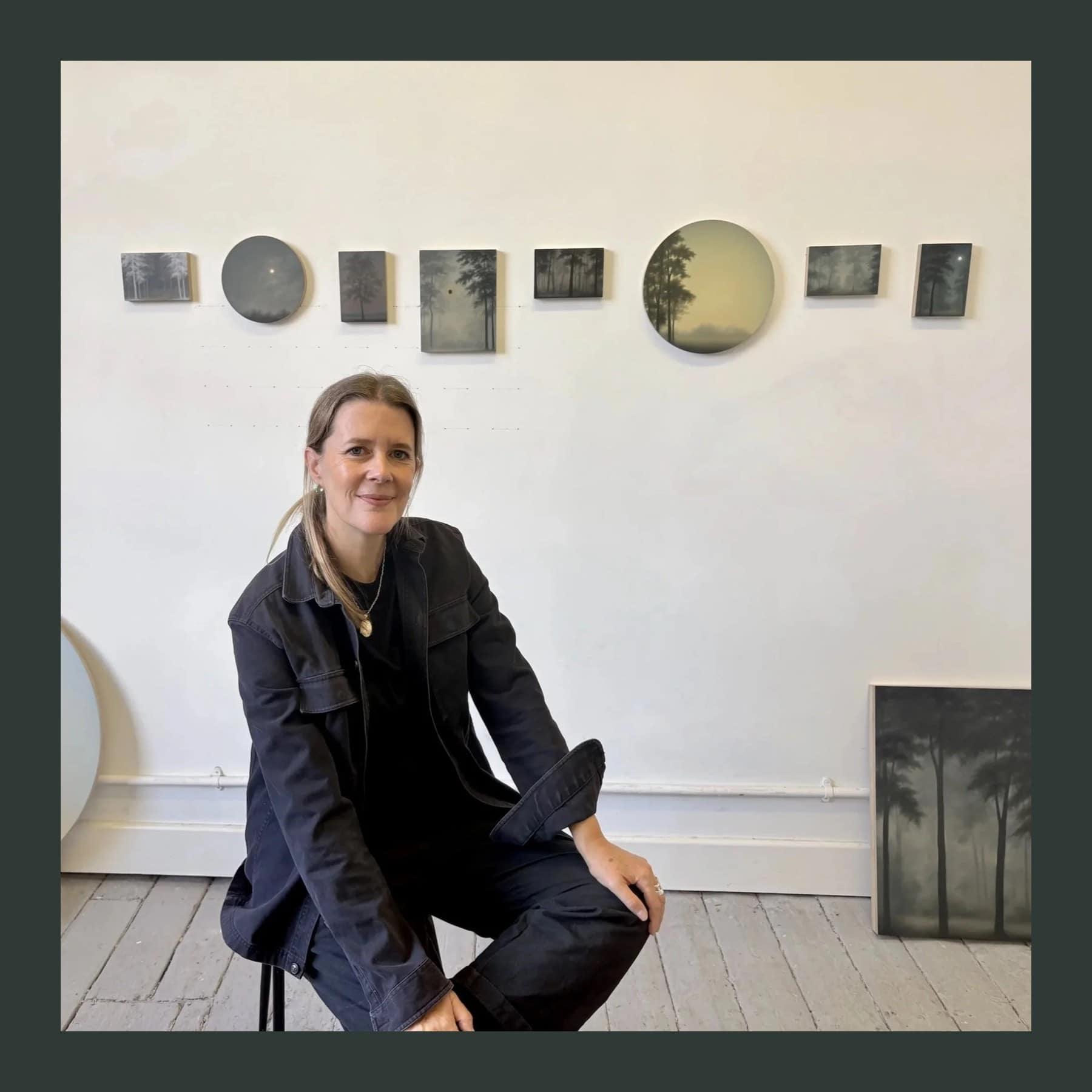 Gill Rocca sitting on a stool in her studio in a black jacket, with a row of small dark landscape paintings and round tondos on the wall behind