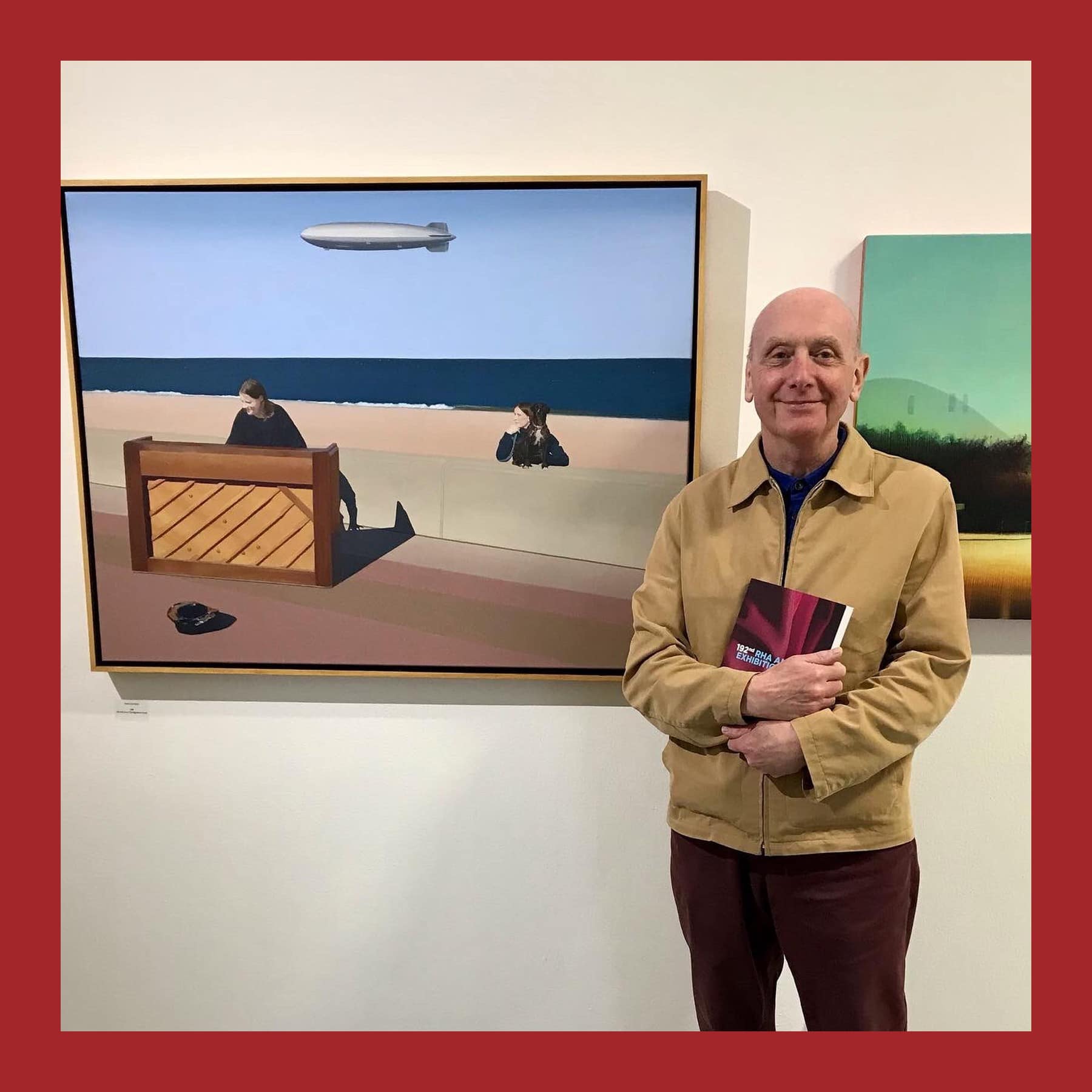 David Hamilton in a tan jacket holding a catalogue beside his painting of a piano on a beach under an airship