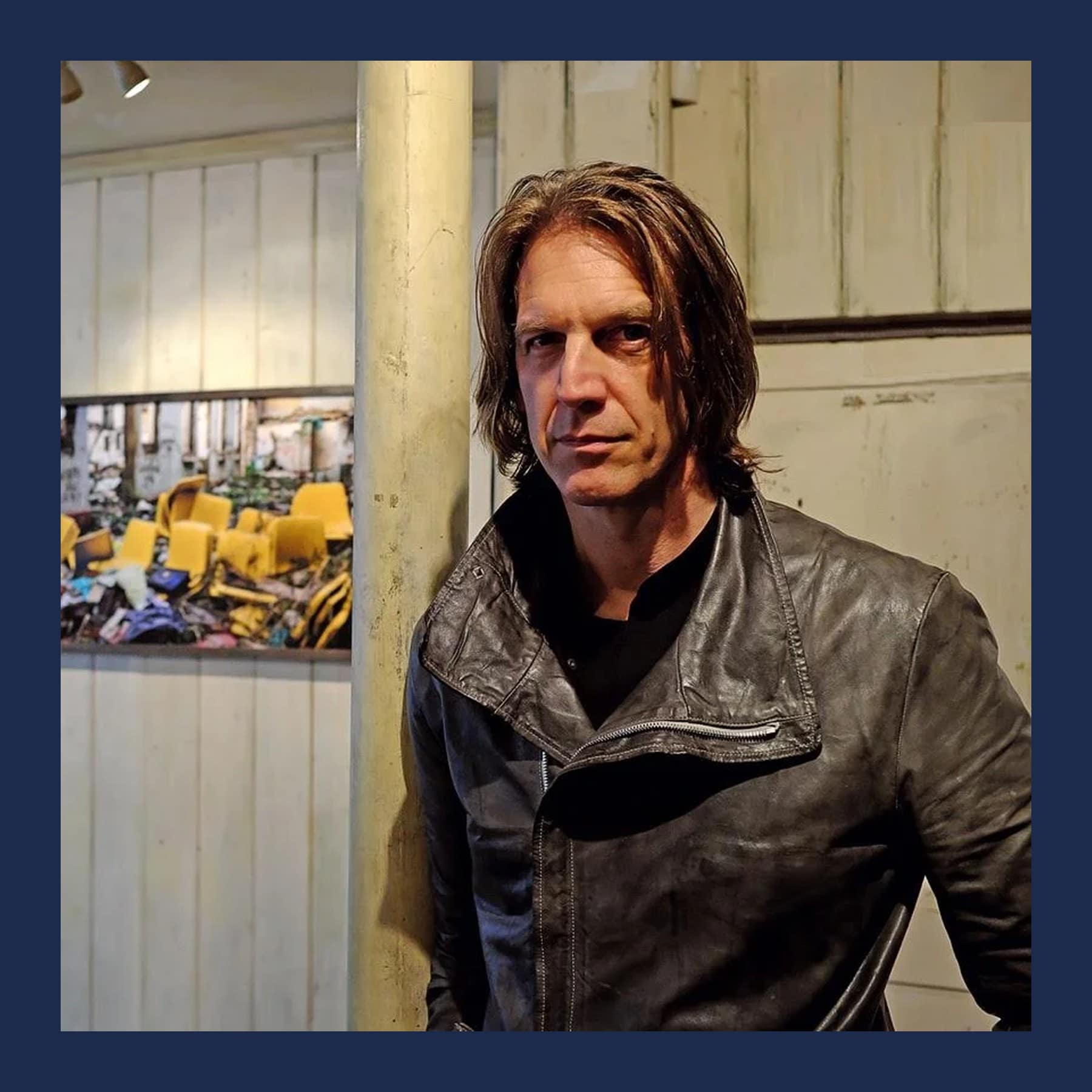 Graham Fink in a black leather jacket standing in a white-panelled gallery beside one of his photographs