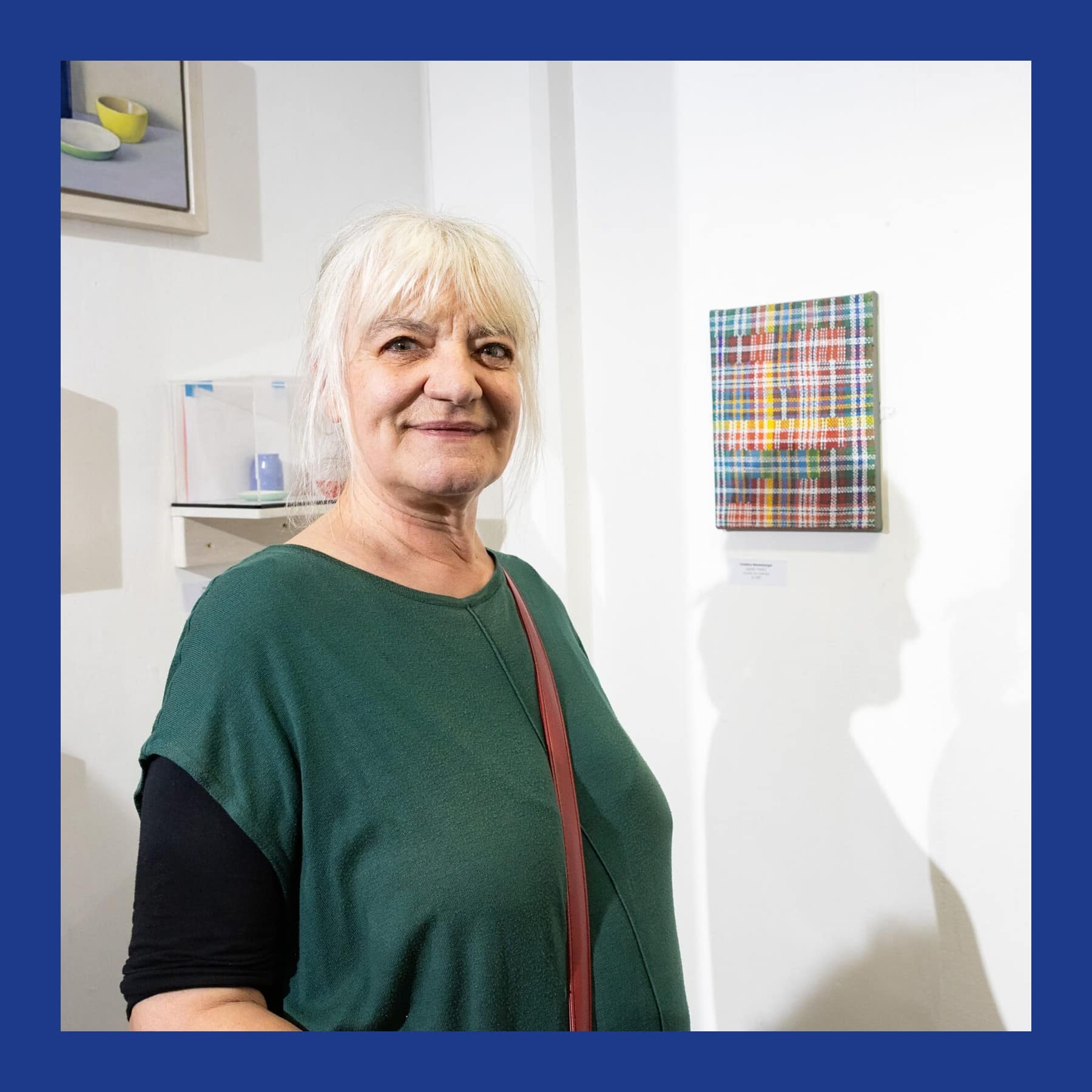 Christina Niederberger, with white hair and a green top, smiling in a gallery beside a small plaid-pattern painting