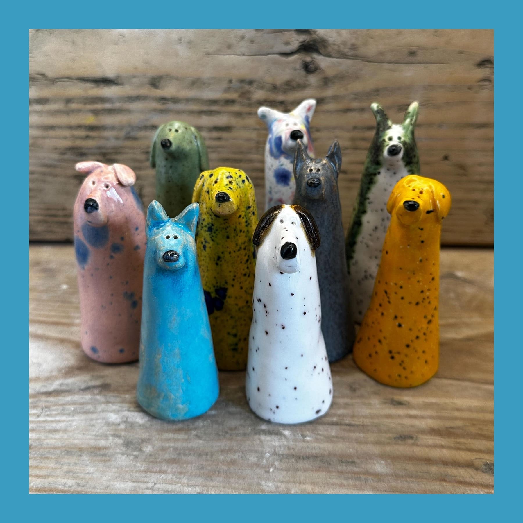 Nine handmade WoodyAndSpud ceramic dogs in pink, blue, yellow, white, grey and orange glazes on a wooden table