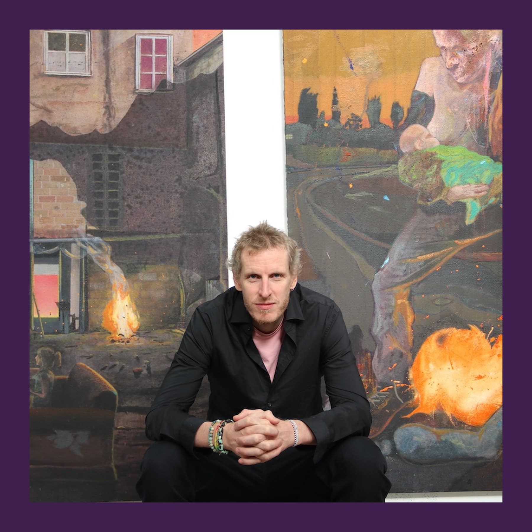 Marcus Cope in a black shirt, crouching in front of two of his large, brightly coloured paintings with fires in them