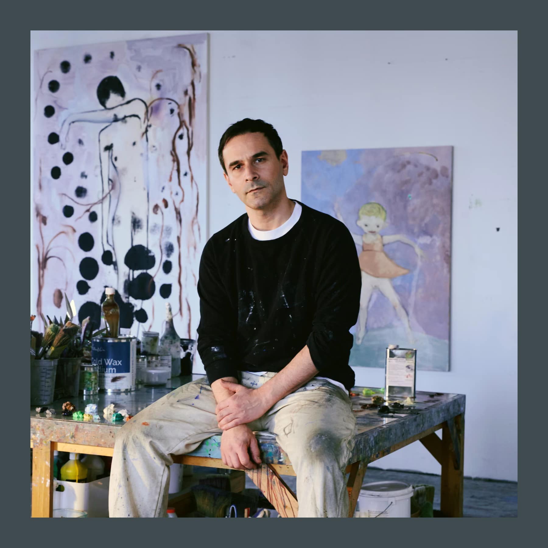 Sanya Kantarovsky in a black jumper sitting on a paint-covered table in his New York studio, with two paintings behind him