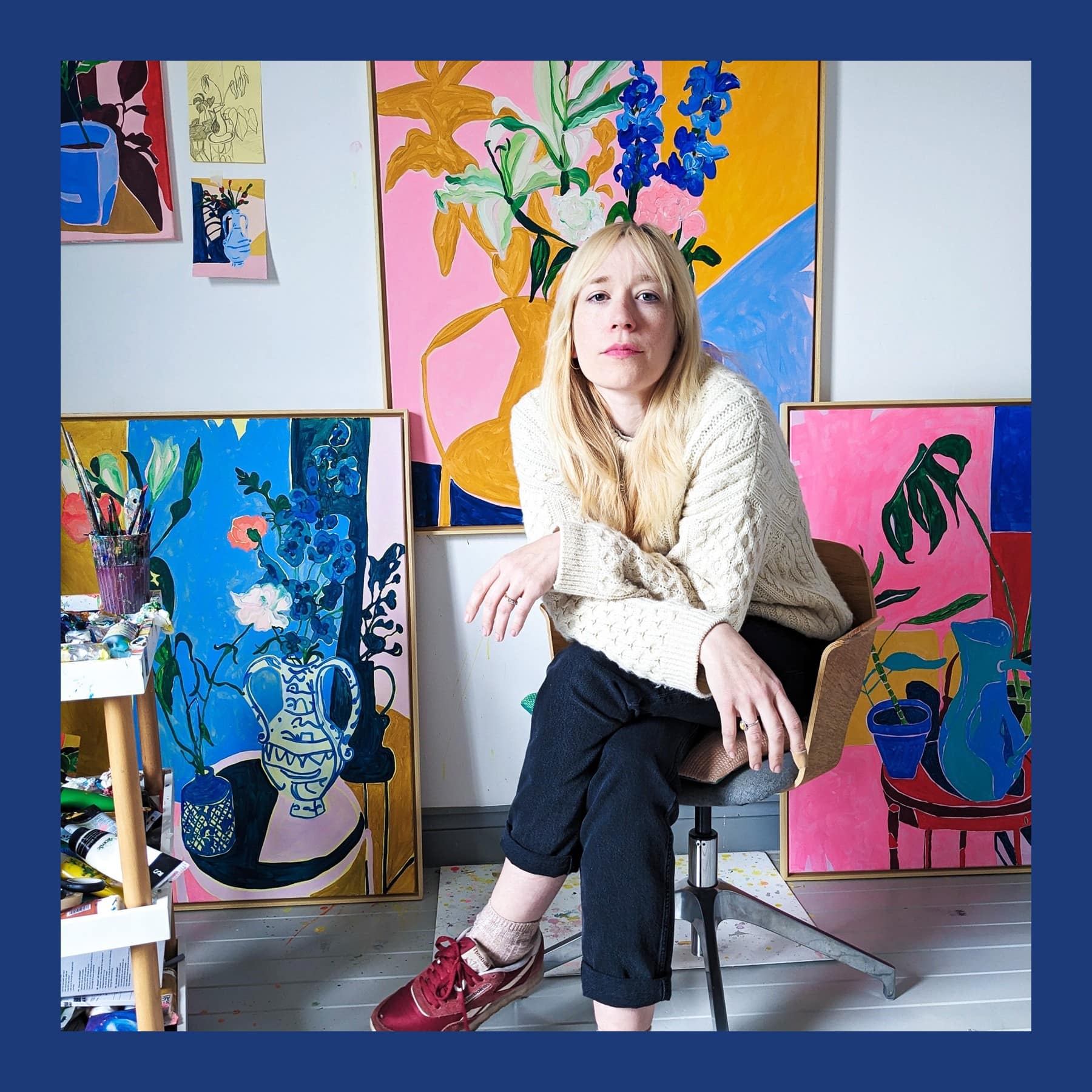Elizabeth Power sitting in her studio in a cream jumper, surrounded by her bright flower and still-life paintings