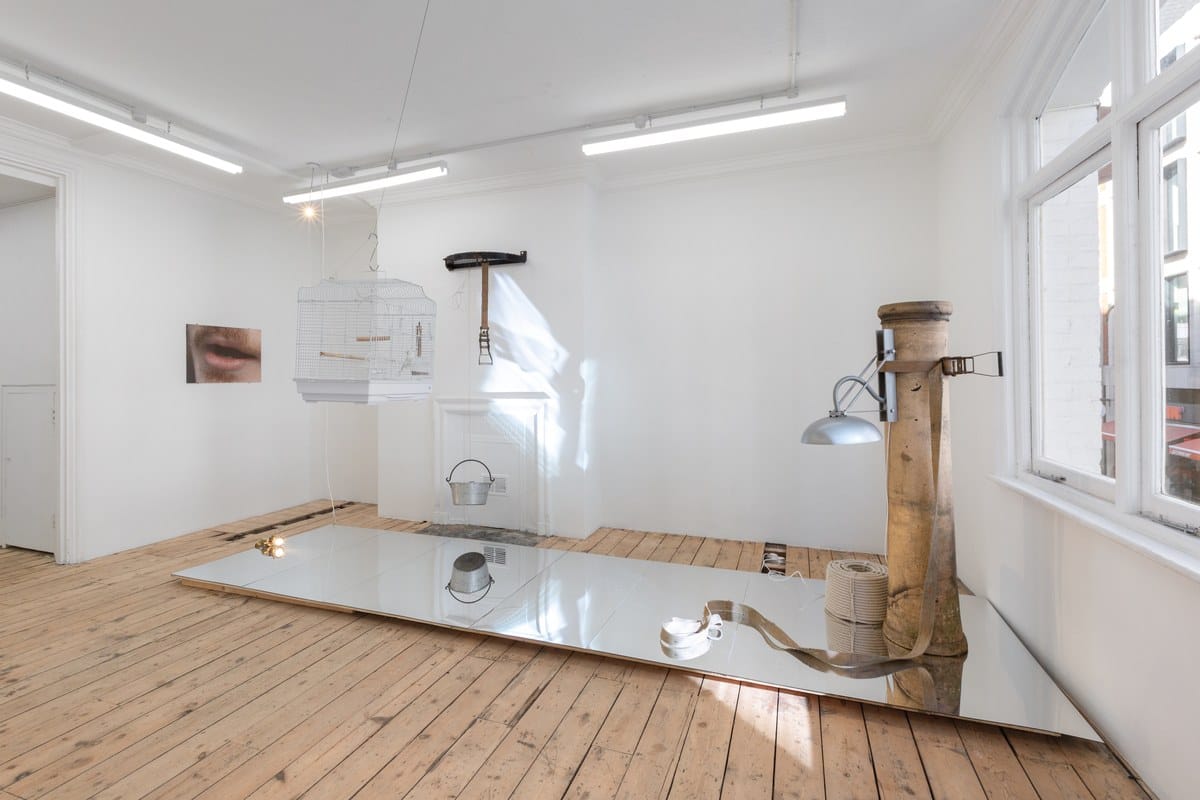 Philip Ewe installation at TINA with a birdcage, a wooden post, lamps and a mirrored platform on a bare floorboard floor