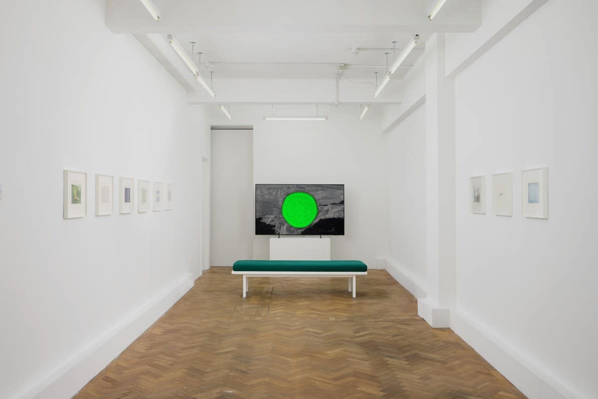 Small framed drawings along two white walls at Arcadia Missa, with a screen and a green bench at the far end