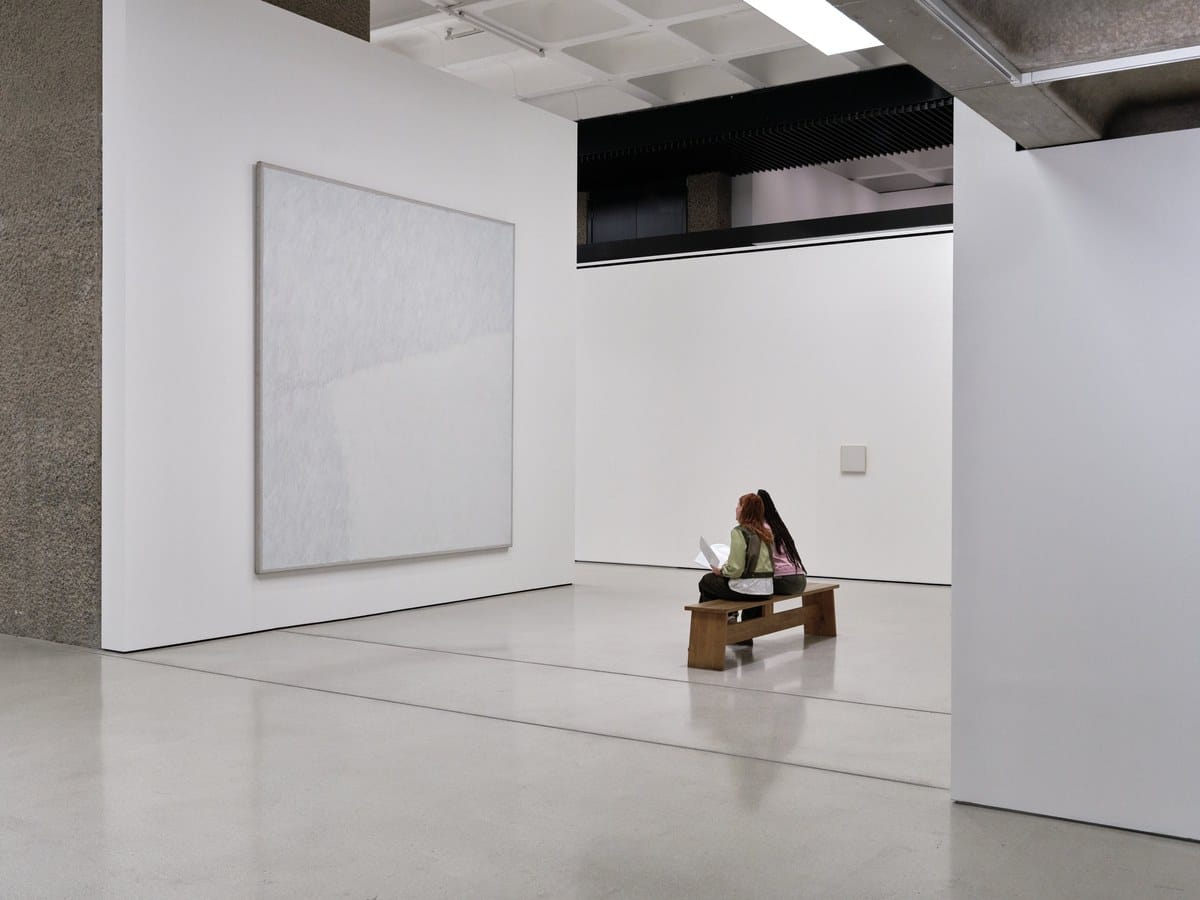 A large white Robert Ryman painting at the Barbican Art Gallery, with a visitor reading on a bench nearby