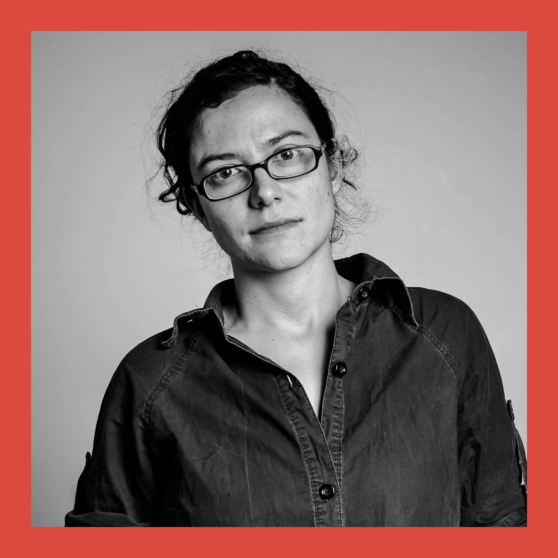 Black and white portrait of Amalia Pica in glasses and a dark denim shirt, head tilted, against a plain wall