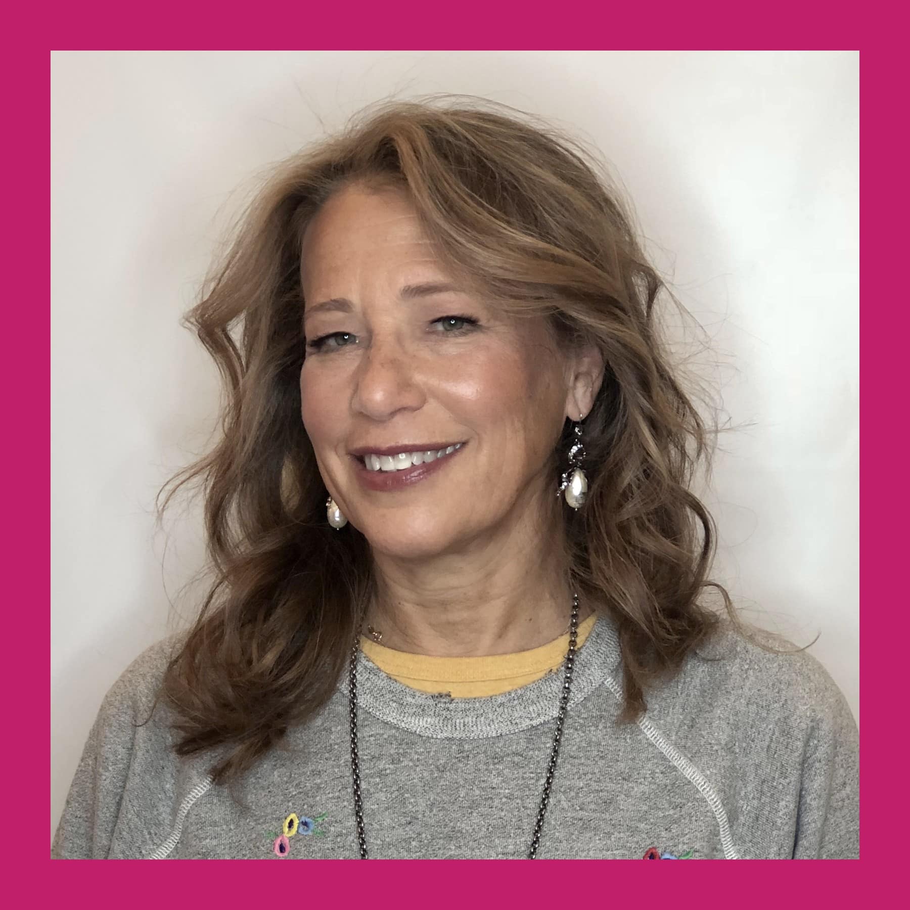 Susan Kare smiling, with wavy shoulder-length hair, pearl earrings and a grey sweatshirt with embroidered flowers