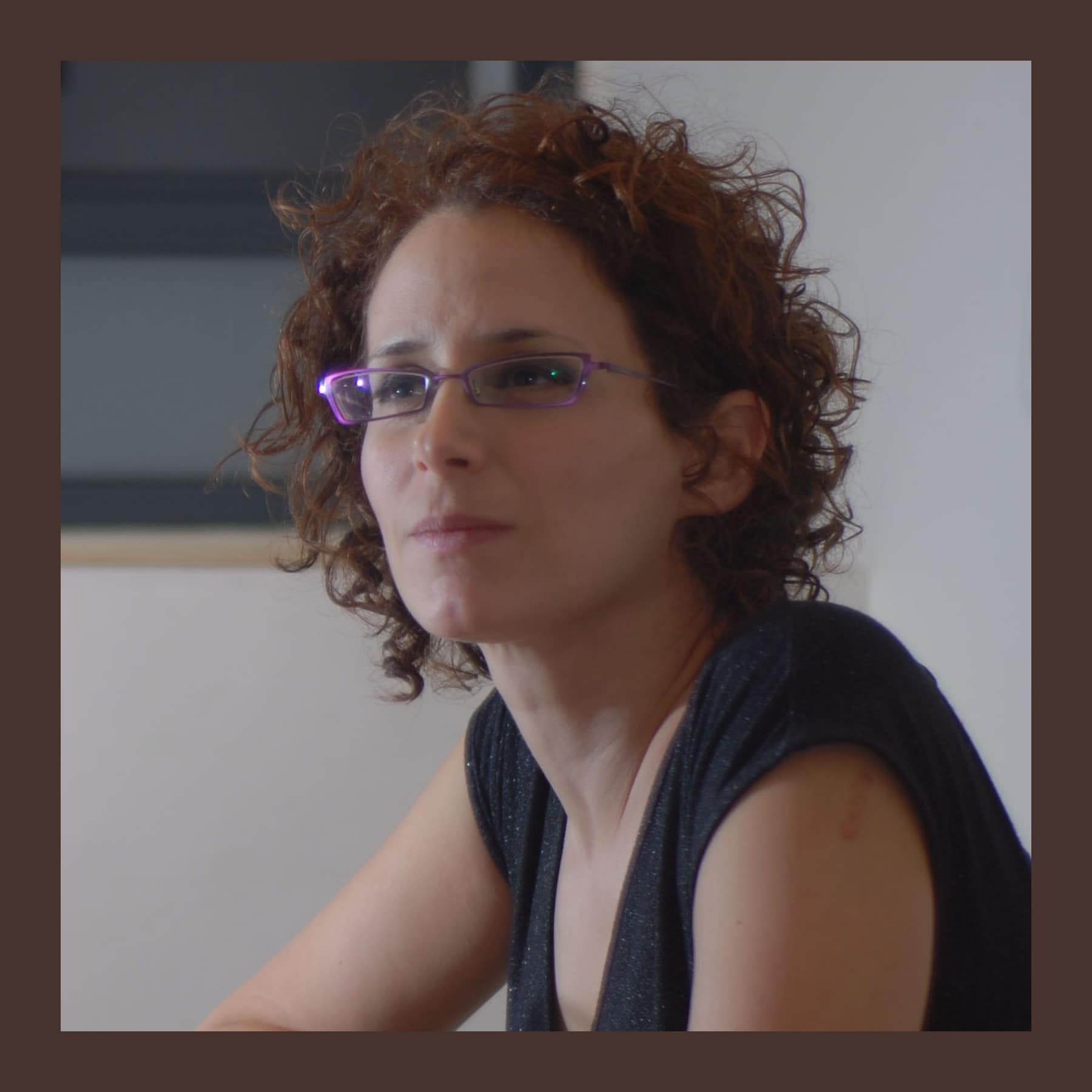 Ronit Baranga with curly auburn hair and purple glasses, seated at a wooden table with her arms folded