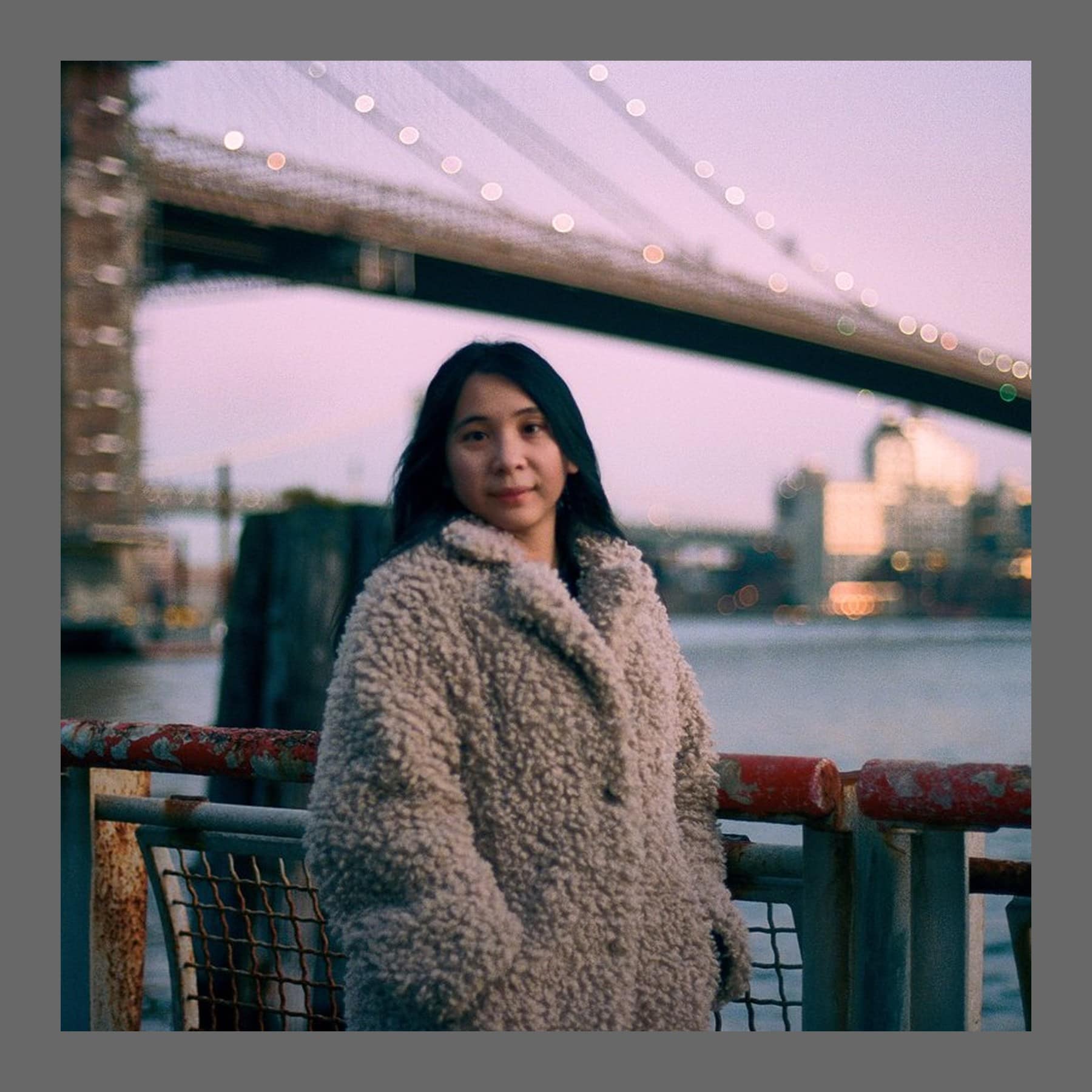 Woman with long dark hair in a cream fleece coat, standing at a railing by the water at dusk with a bridge behind