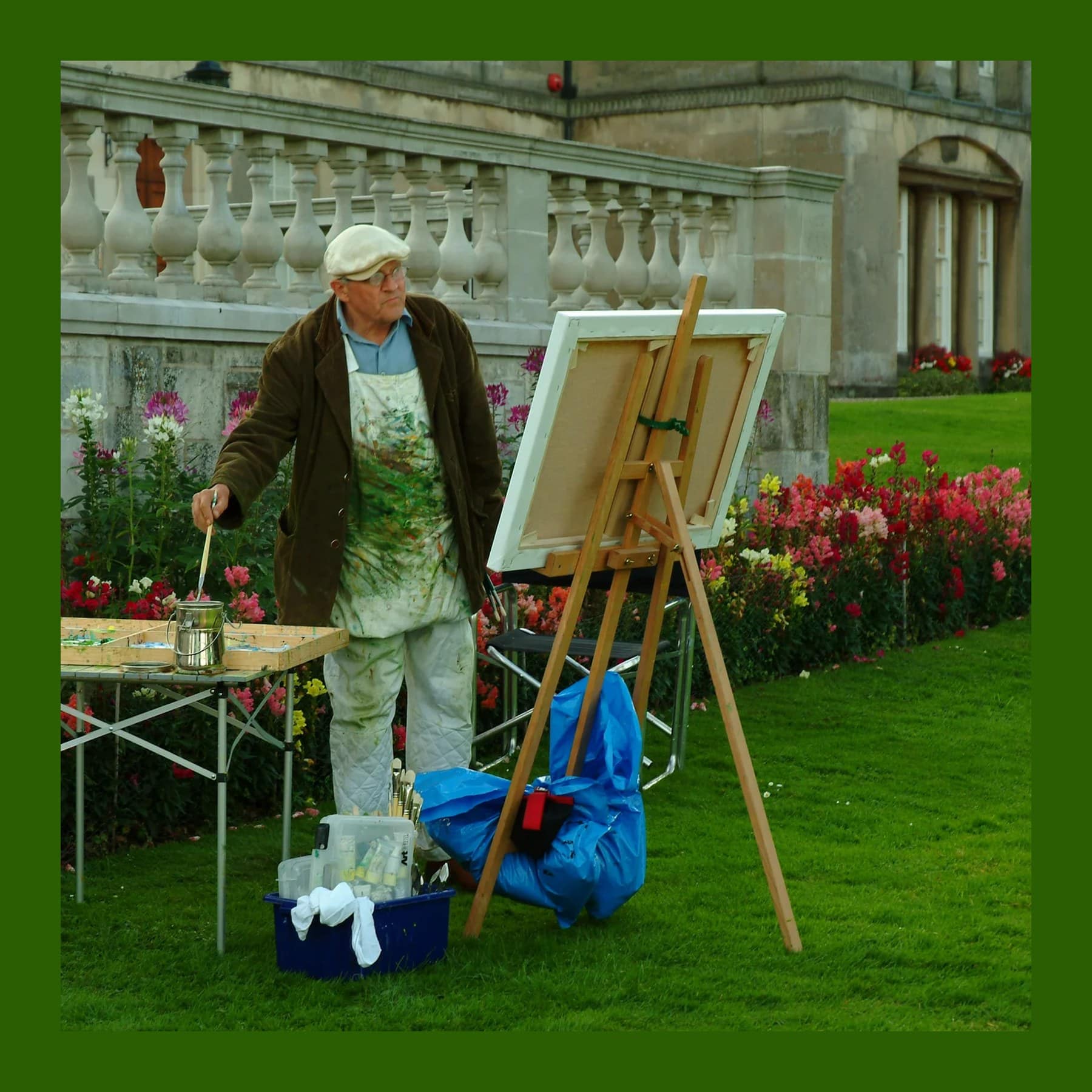 David Hockney in a flat cap and paint-spattered apron painting at a wooden easel on a lawn beside flowerbeds