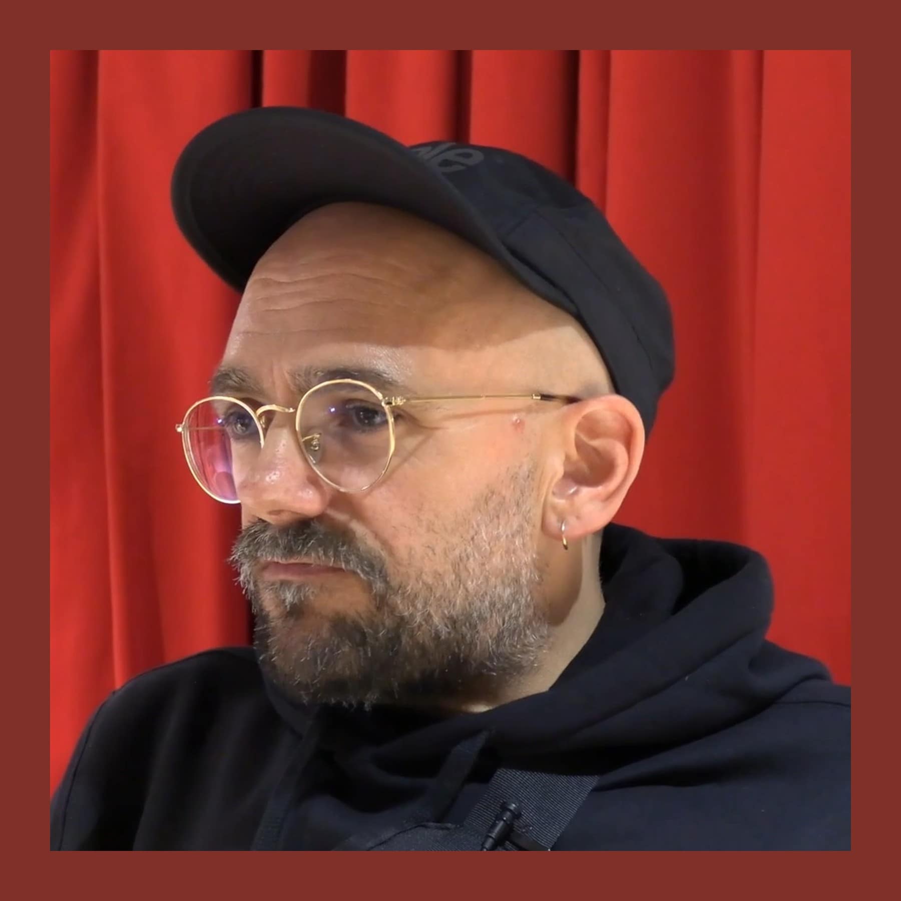 Ryan Gander in a black cap and round glasses, with a grey beard, in front of a red curtain