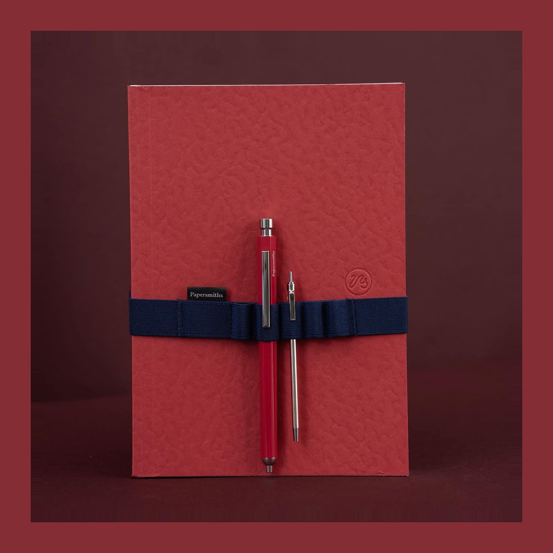 Papersmiths Sorrel Red A5 notebook with a navy elastic band holding a red Primo Pen and a steel Pocket Pen