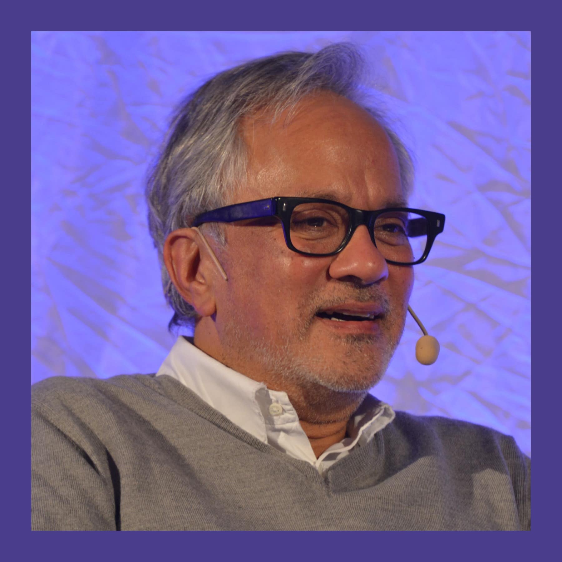 Anish Kapoor in 2017, wearing dark-framed glasses and a grey jumper, speaking with a headset microphone