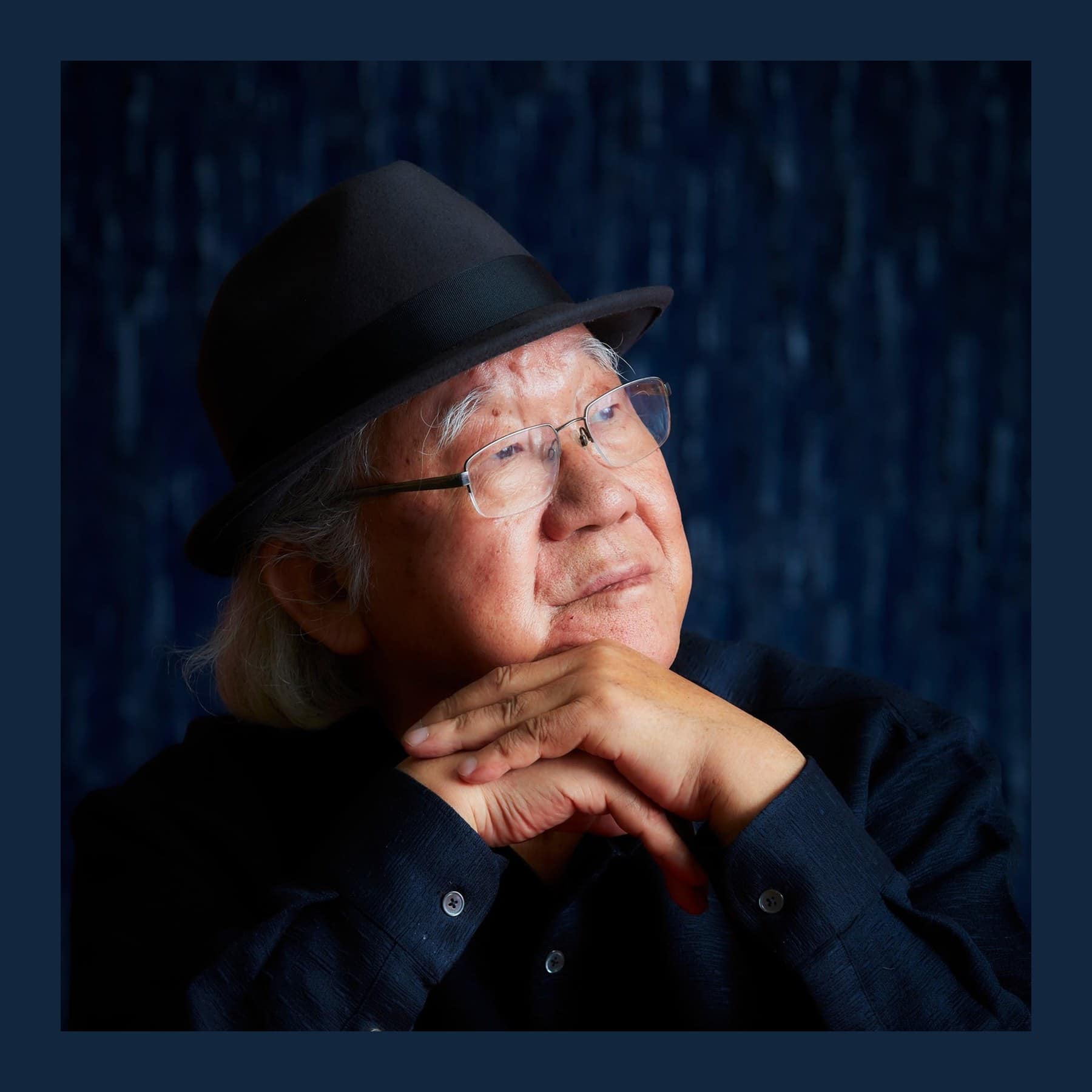 Ha Chong-Hyun in a black trilby and rimless glasses, chin resting on his hands, against a dark blue background