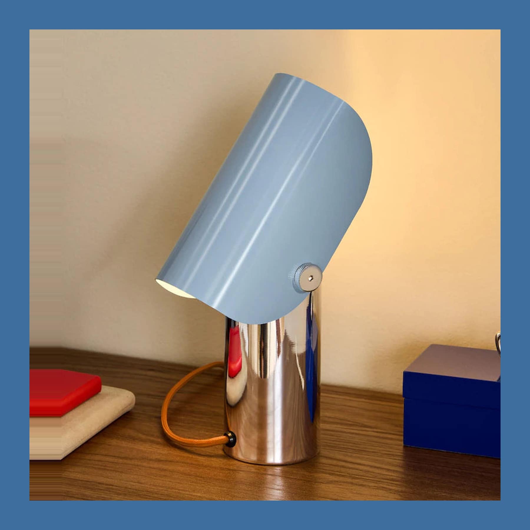 MADE x Barbican Curve Table Lamp in Chrome and Blue lit on a bedside table, its tubular shade tilted forward on a polished chrome base