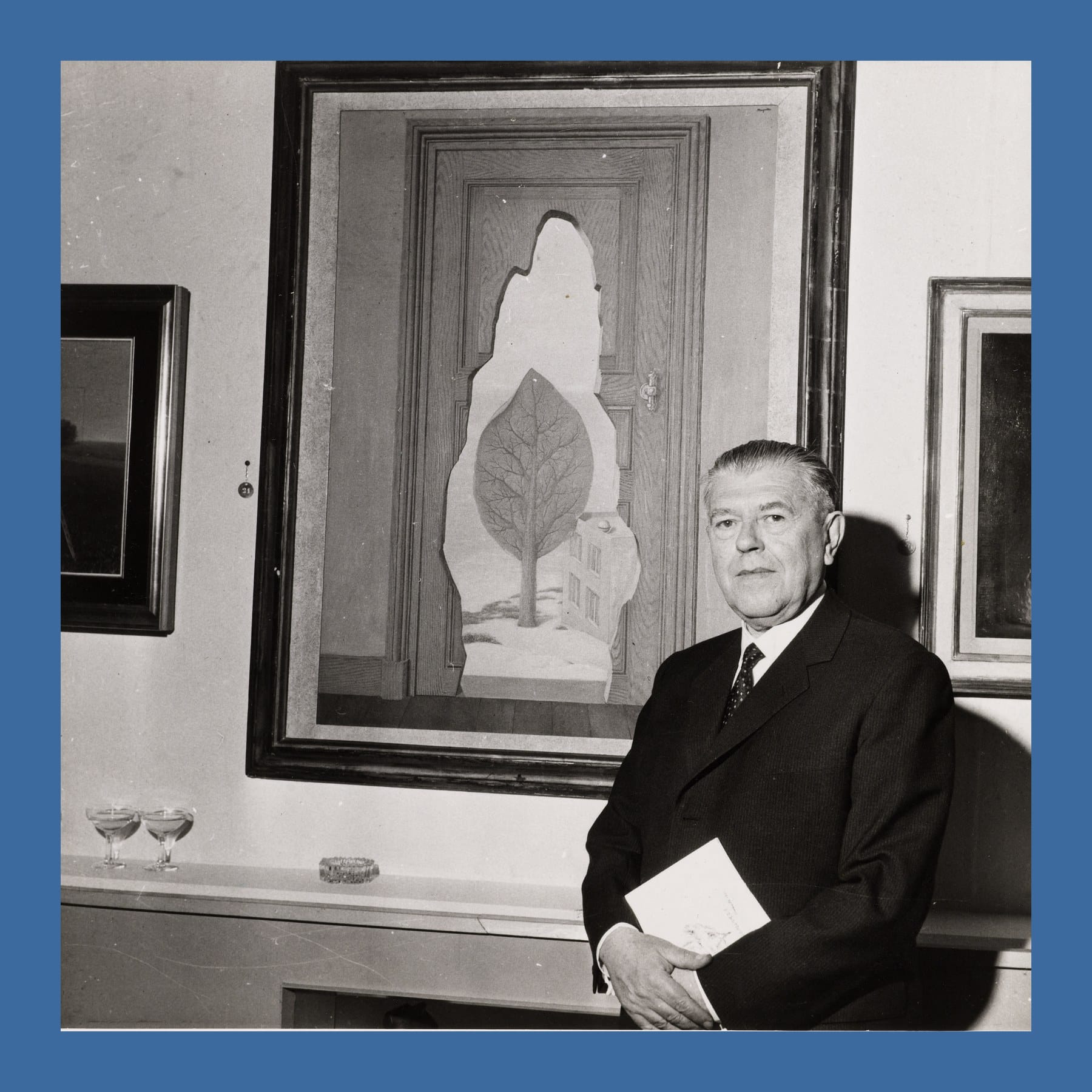 Black and white photograph of René Magritte in a dark suit standing in front of one of his framed paintings