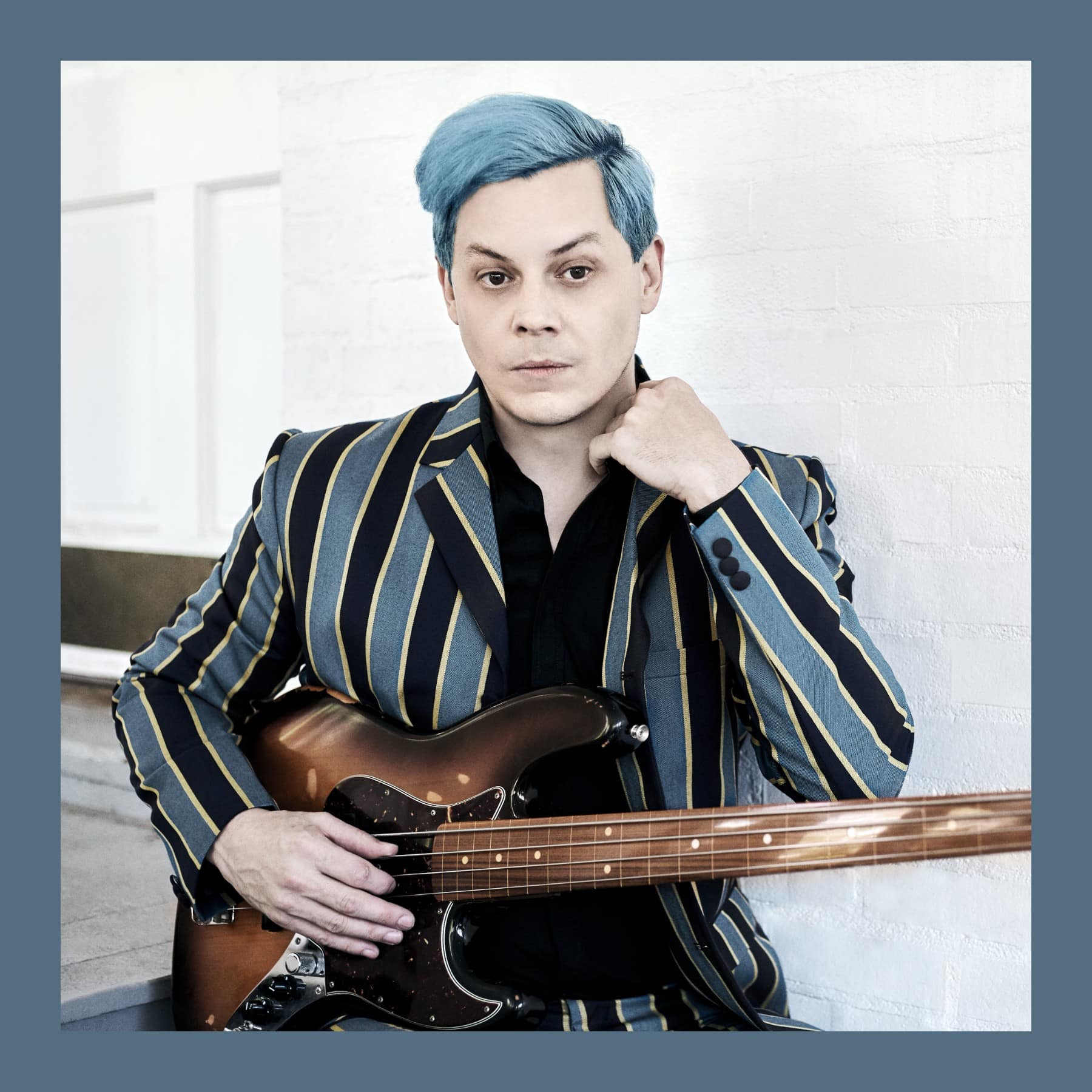 Jack White with blue hair in a striped blazer, holding a bass guitar against a white brick wall