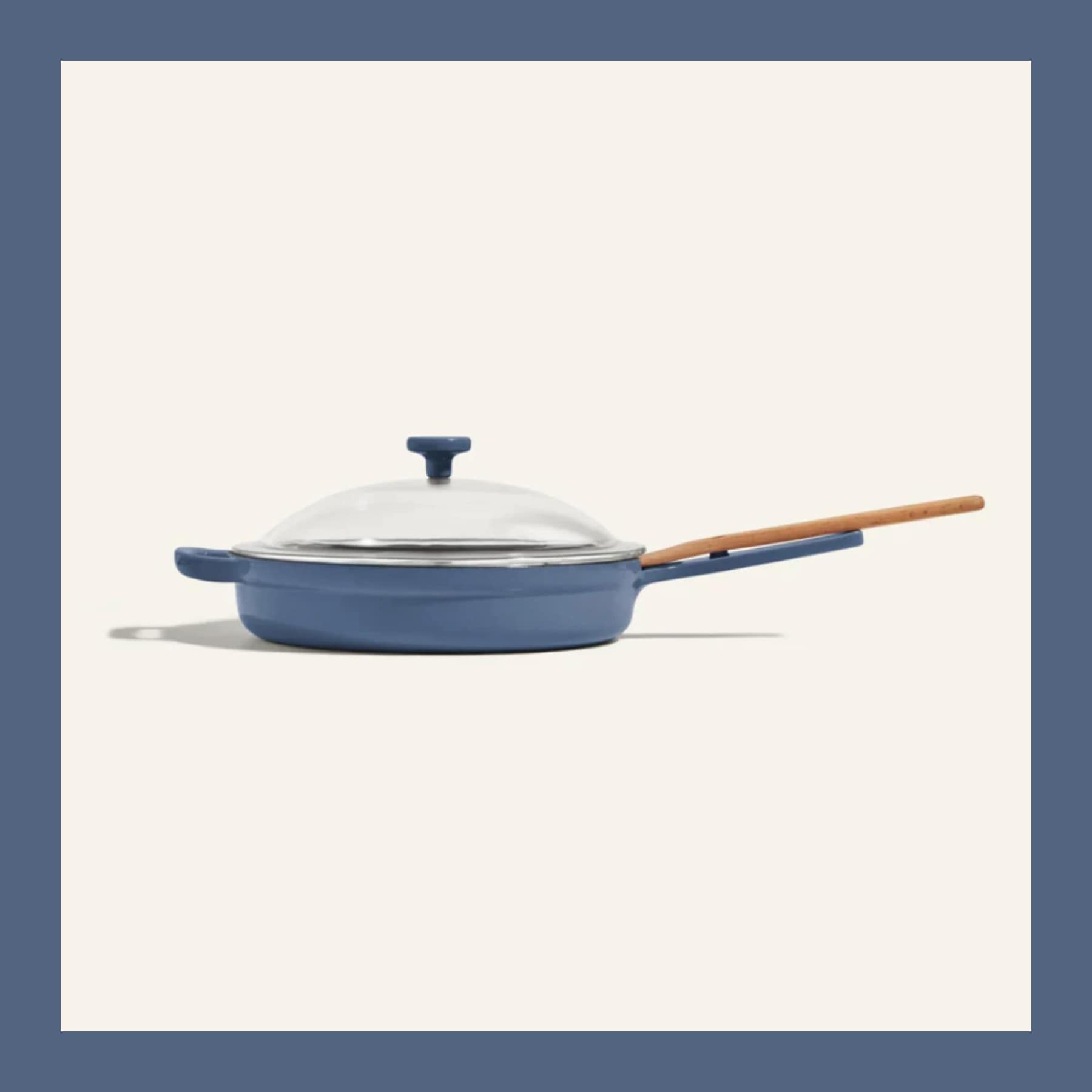 The Our Place Always Pan 2.0 in Blue Salt with its glass lid and beechwood spatula, on a cream ground inside a blue border.