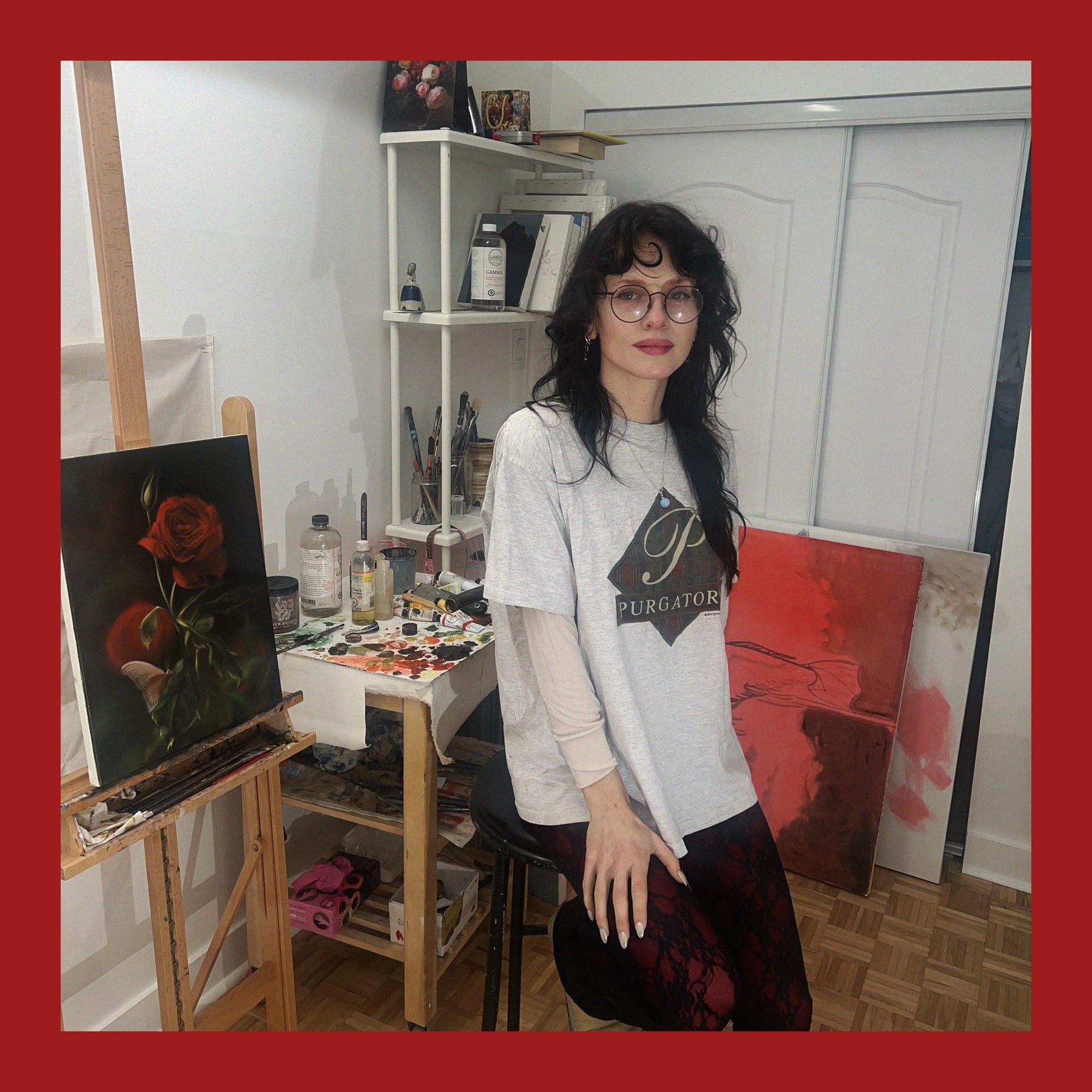 Rebecca Storm seated on a stool in her Montreal studio beside an easel with a painting of red roses