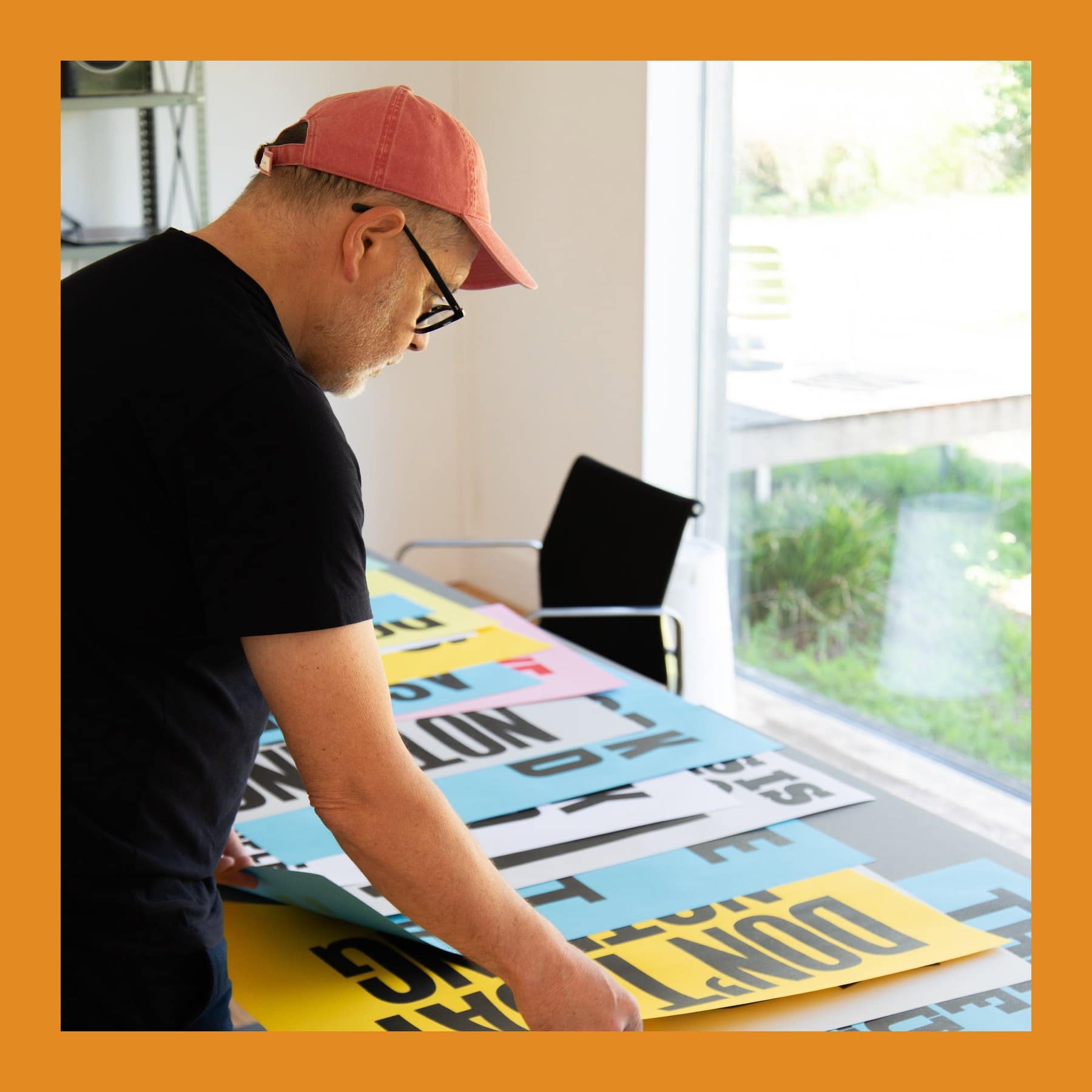 Anthony Burrill in a red cap and glasses laying out colourful letterpress posters on a long table in his studio