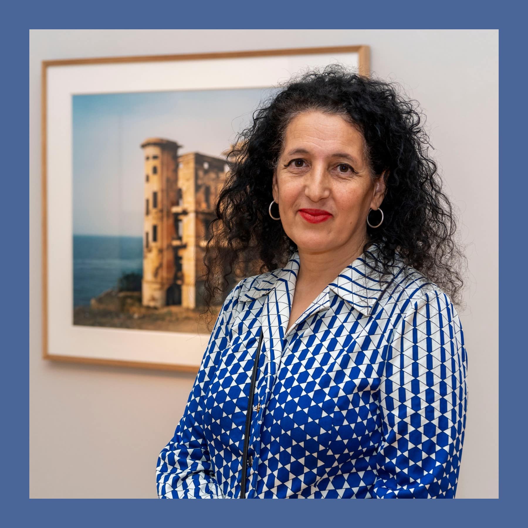Zineb Sedira with curly dark hair and a blue and white patterned shirt, standing beside one of her framed photographs