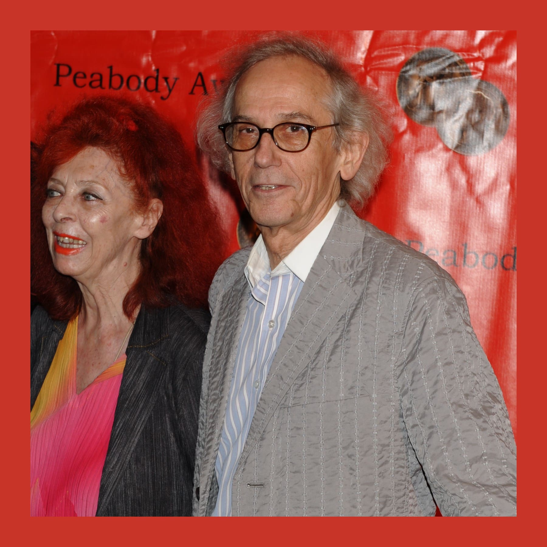Christo in glasses and a grey jacket beside Jeanne-Claude with red hair, in front of a red backdrop