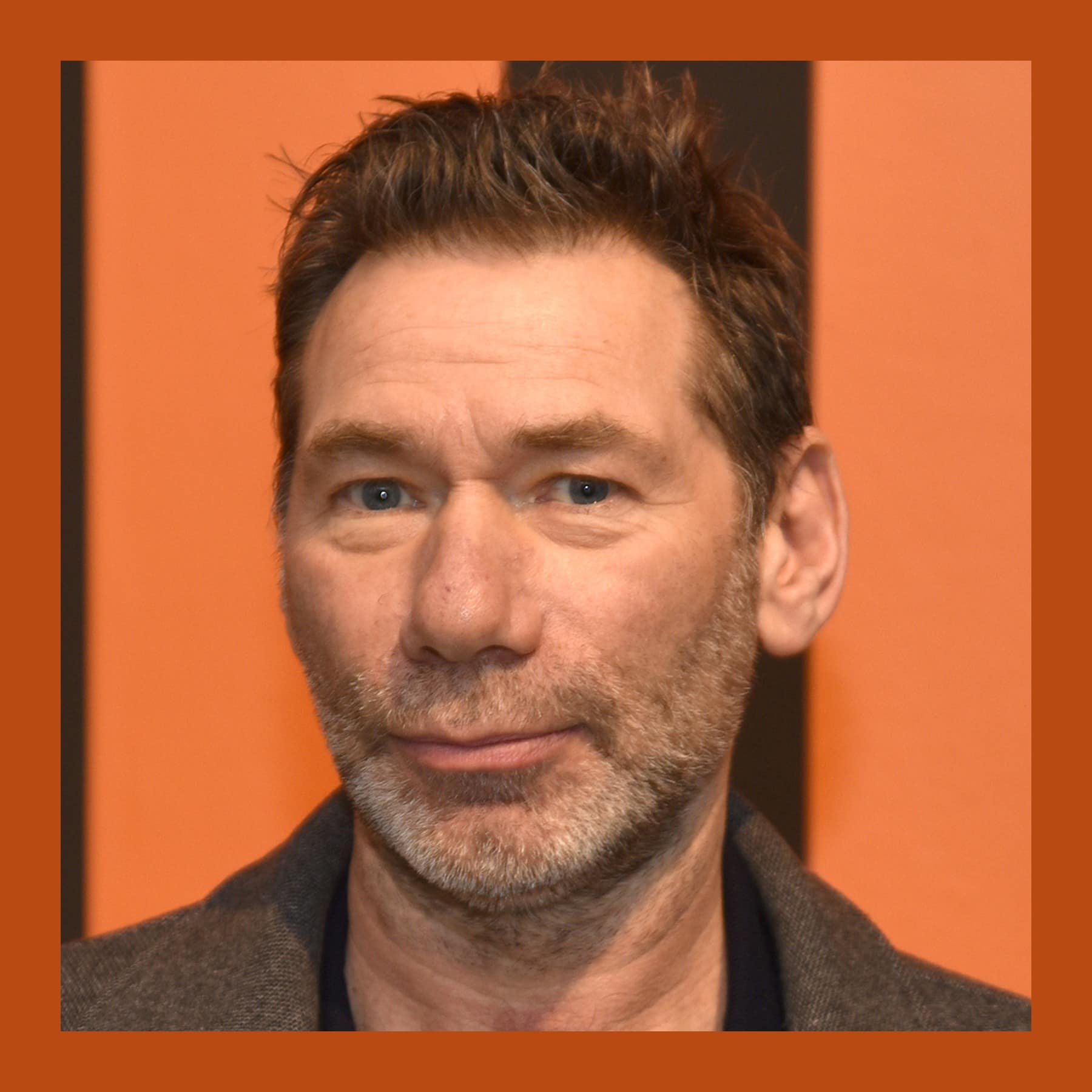 Mat Collishaw, with short brown hair and stubble, in front of an orange wall