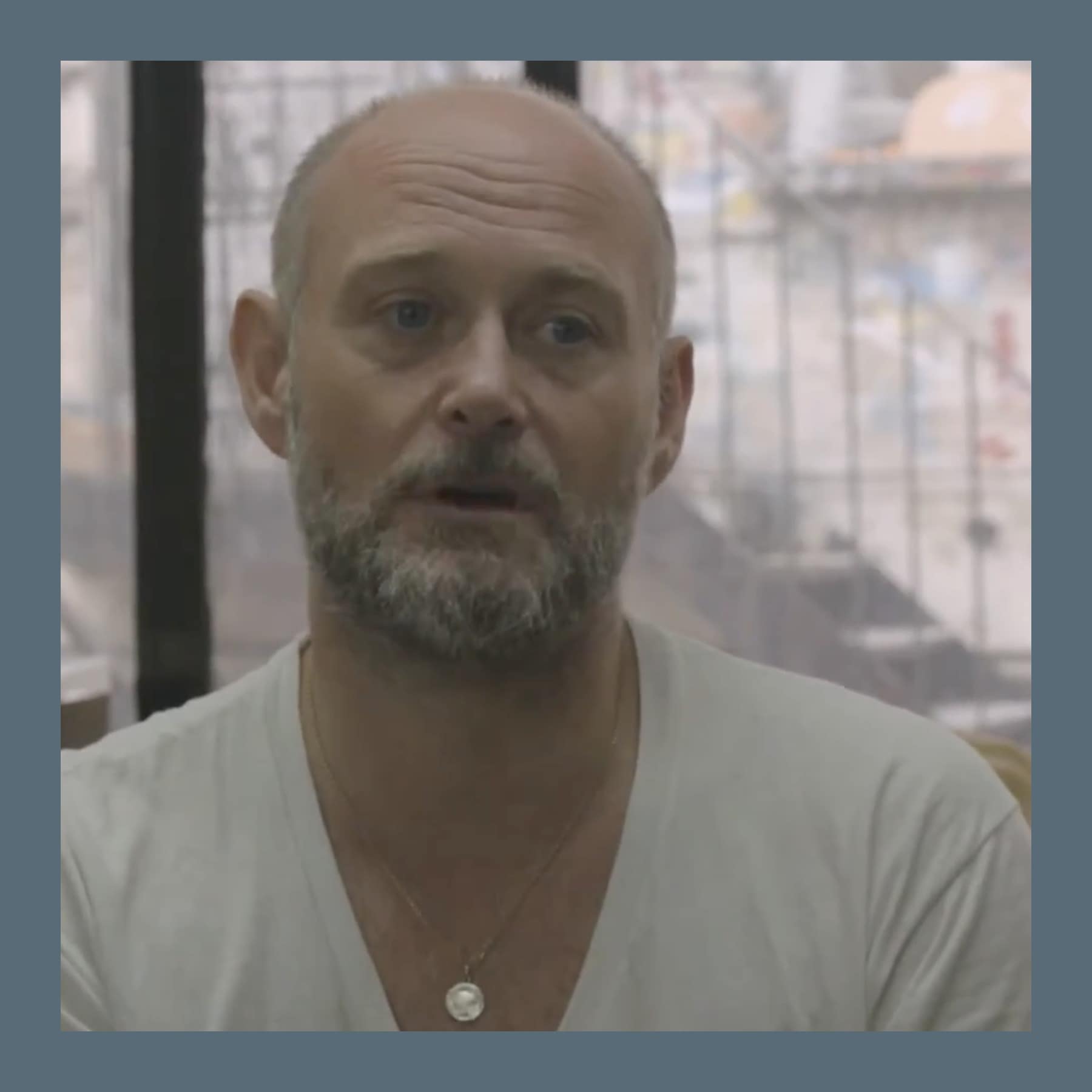 Steven Claydon, bald with a grey beard, in a pale V-neck T-shirt, speaking to camera