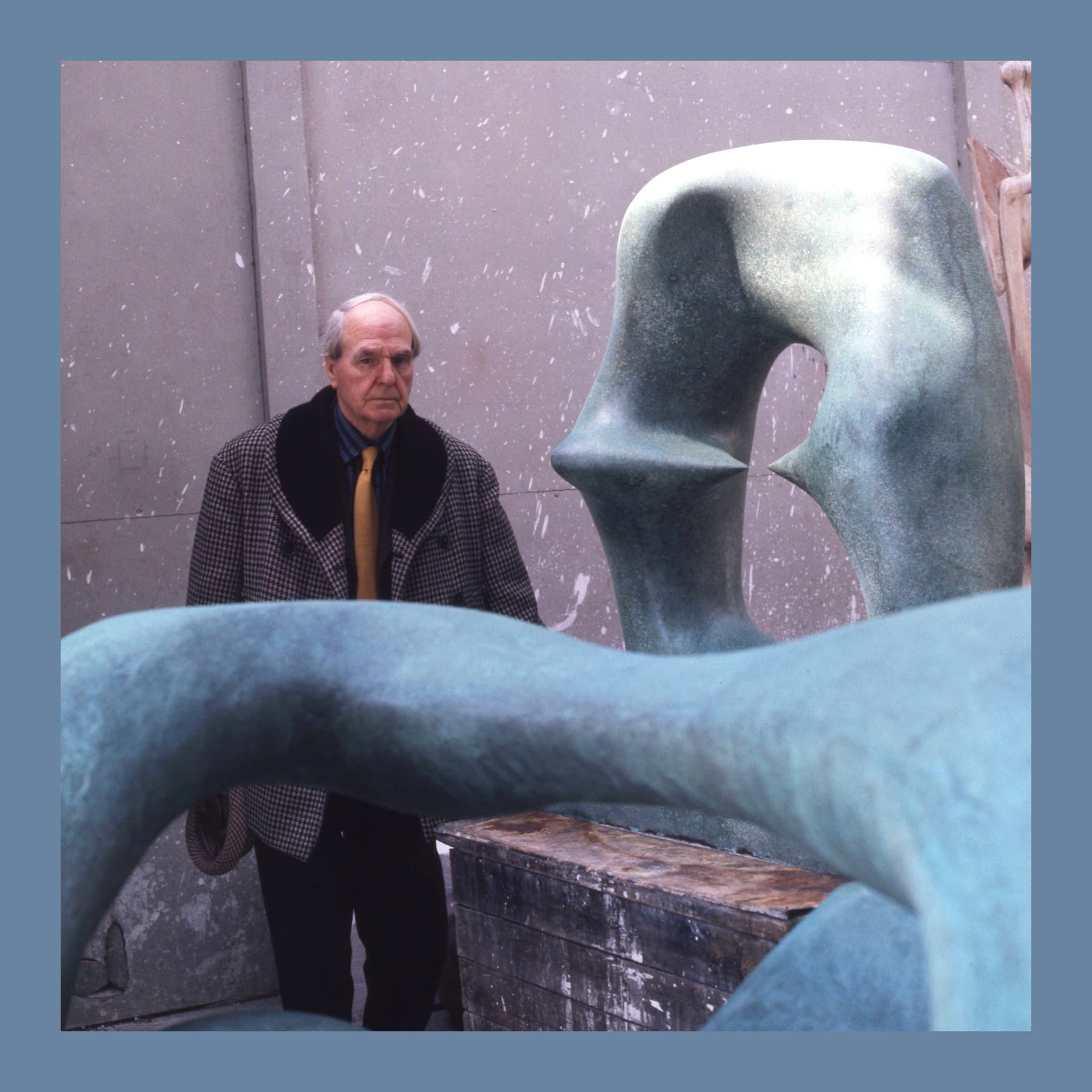 Henry Moore in a tweed coat and yellow tie standing behind large green bronze sculptures in his studio
