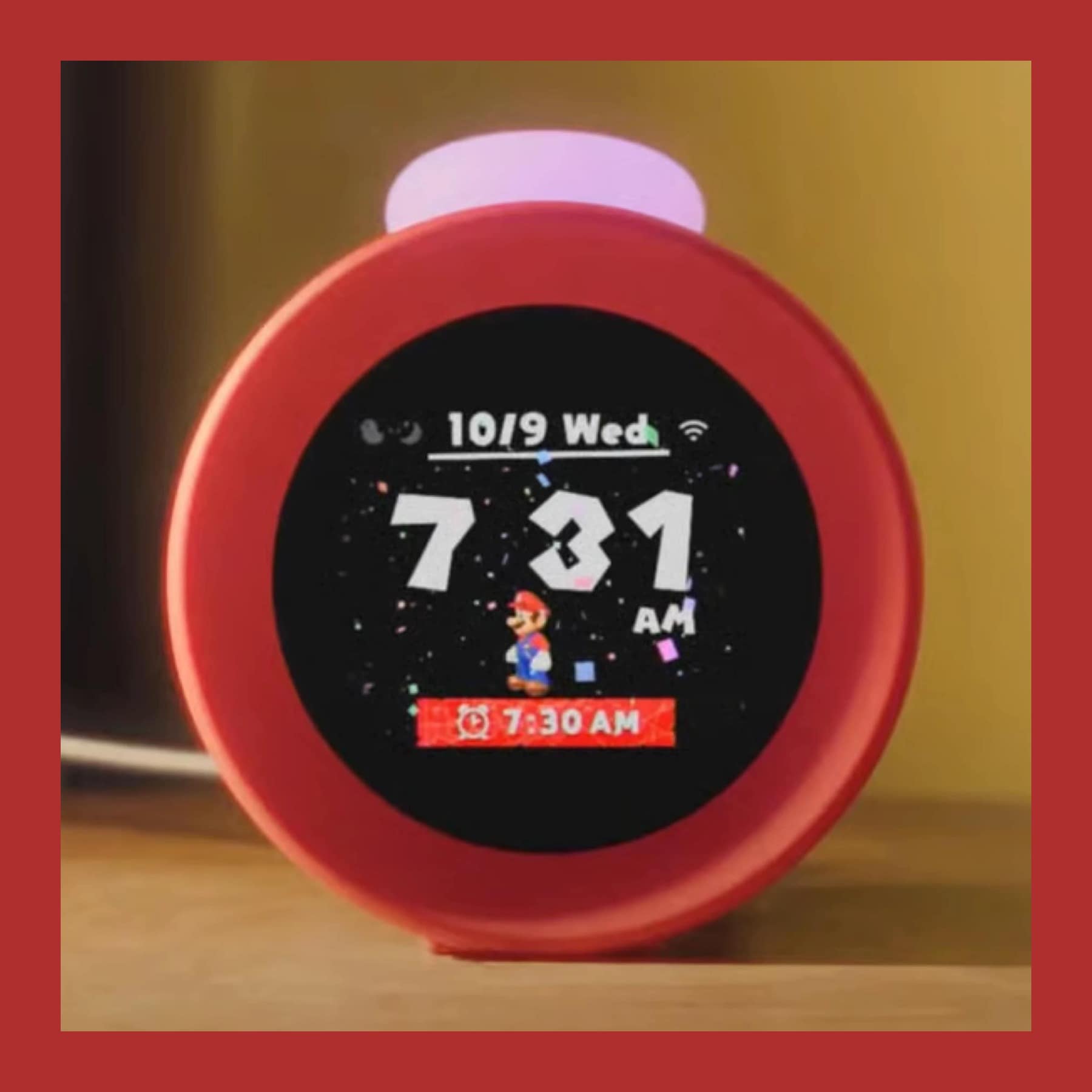 A red Nintendo Alarmo clock on a bedside table, its screen showing Mario and a 7:30 alarm, inside a red border.