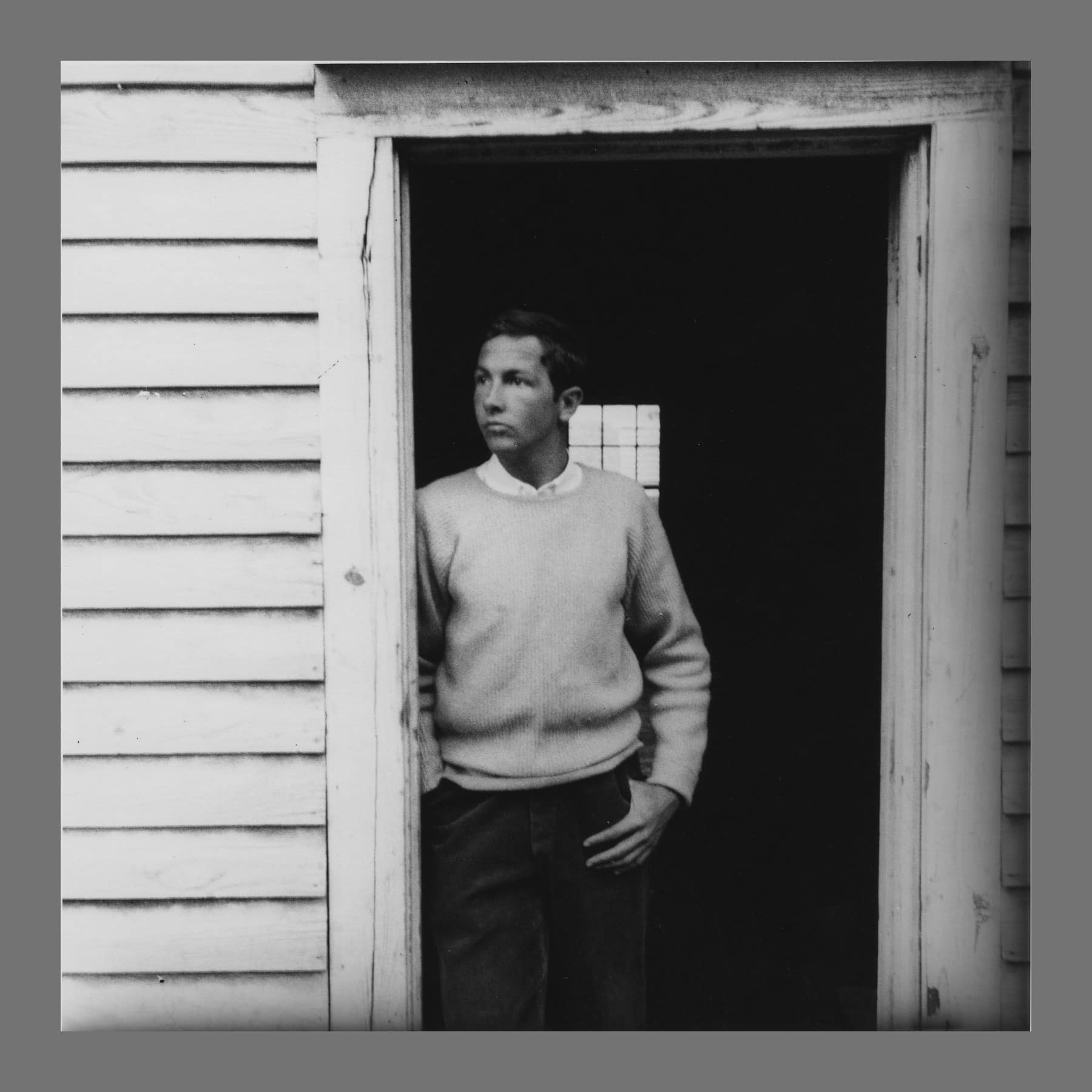 Black-and-white photo of a young man in a pale jumper leaning in the doorway of a wooden building
