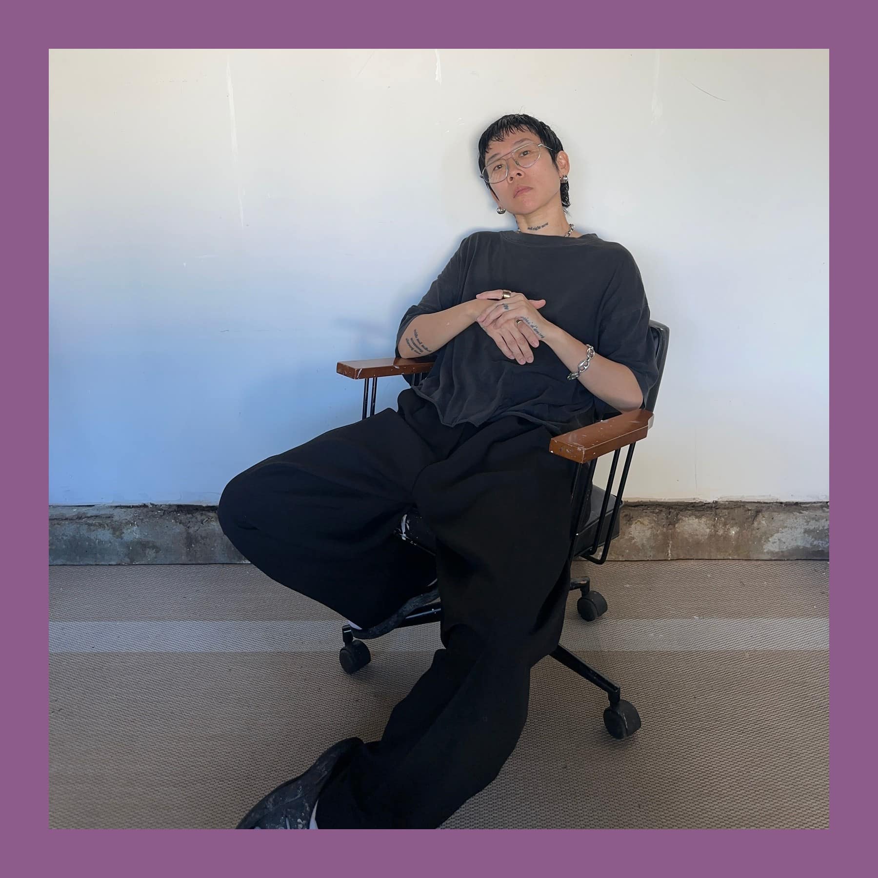 Michelle Jane Lee leaning back in an office chair against a white wall, in round glasses and dark clothes