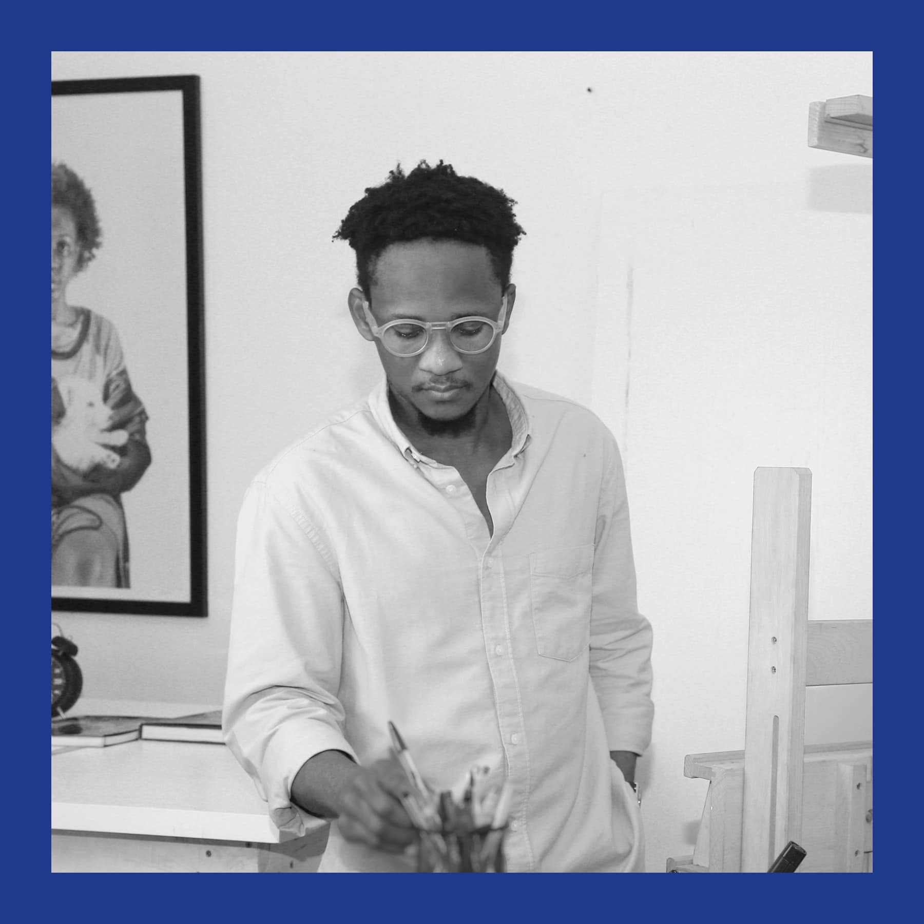 Black and white photo of Oscar Ukonu in glasses and a light shirt, reaching into a pen pot at his studio desk