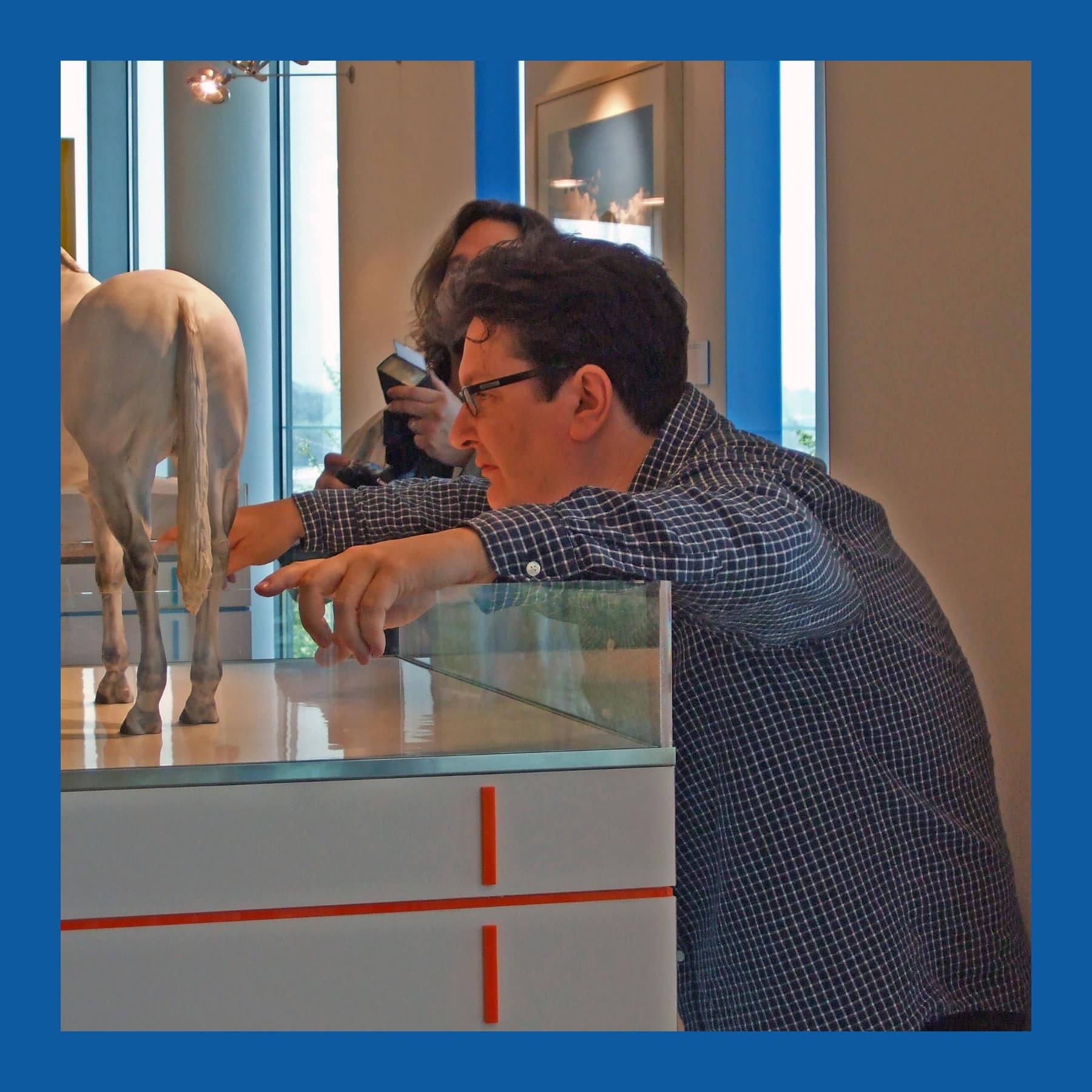 Mark Wallinger in glasses and a checked shirt leaning over a glass case to point at a model of a white horse