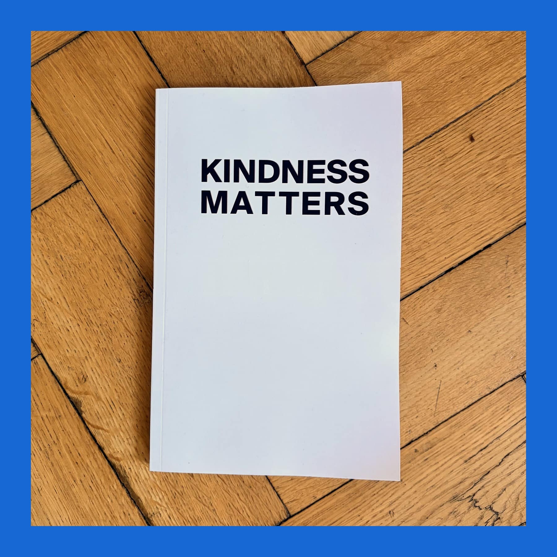 White KINDNESS MATTERS paperback notebook with bold black title lettering, lying on an oak parquet floor
