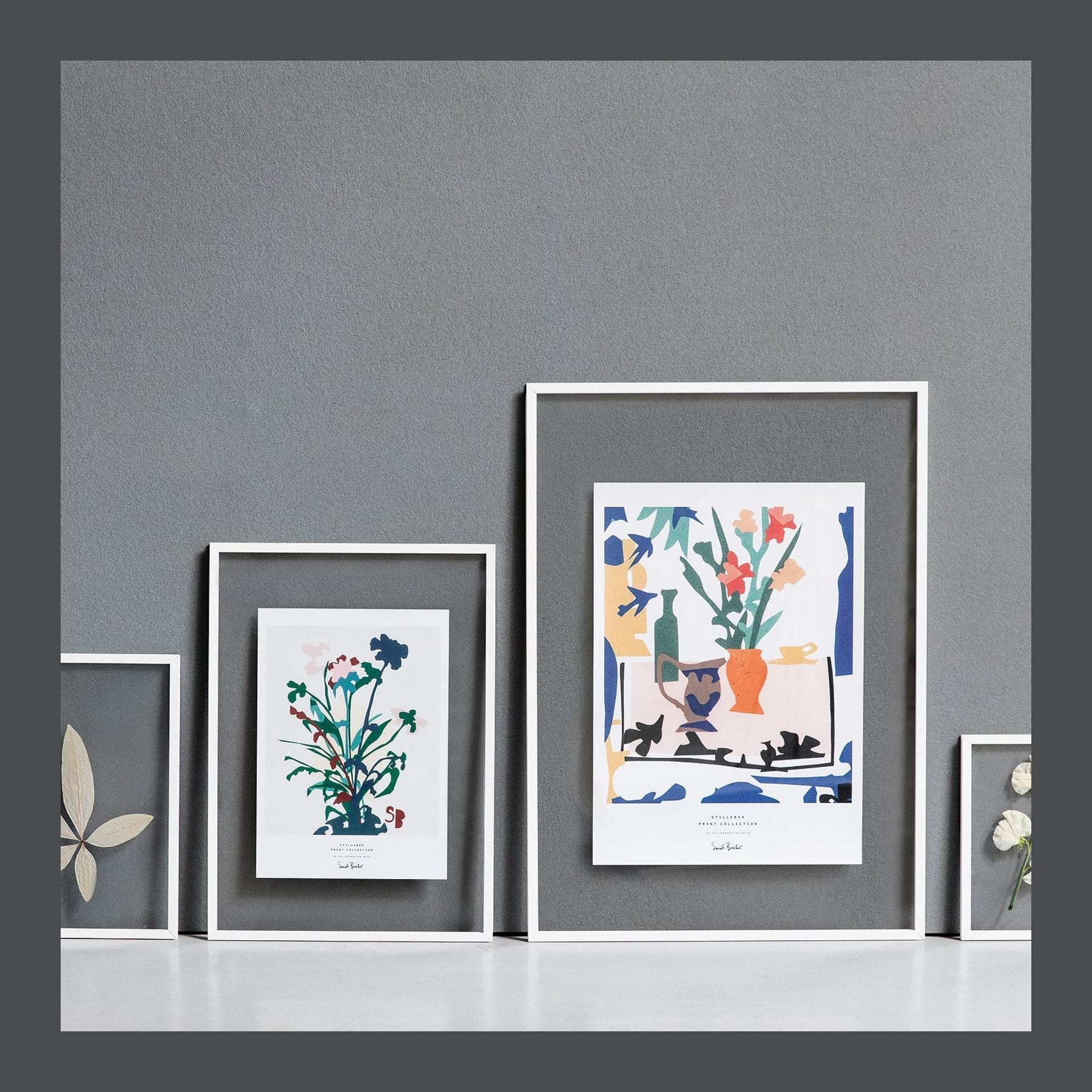 White Moebe frames leaning on a shelf against a grey wall, holding pressed plants and a colourful print