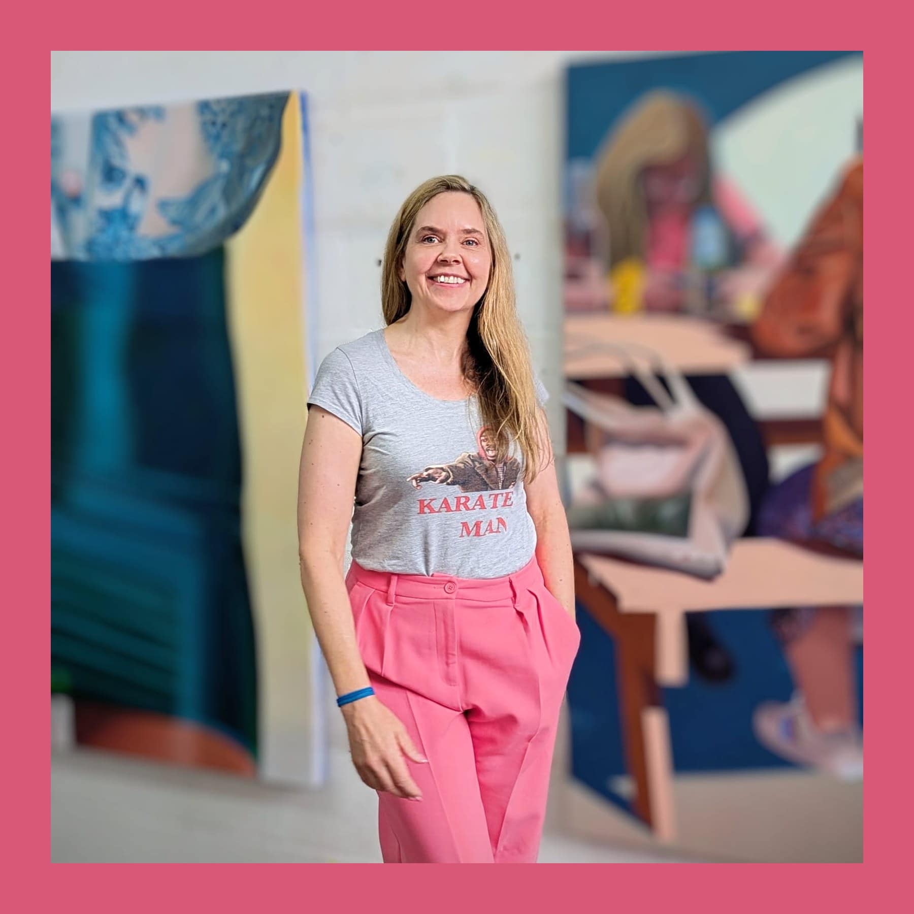 Sally Kindberg smiling in a grey T-shirt and pink trousers in her studio, with her paintings behind her