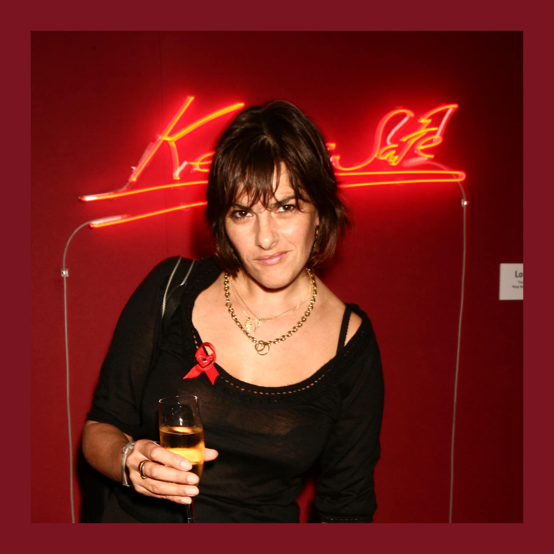 Tracey Emin in a black dress with a red ribbon, holding a glass of wine in front of a red neon on a red wall
