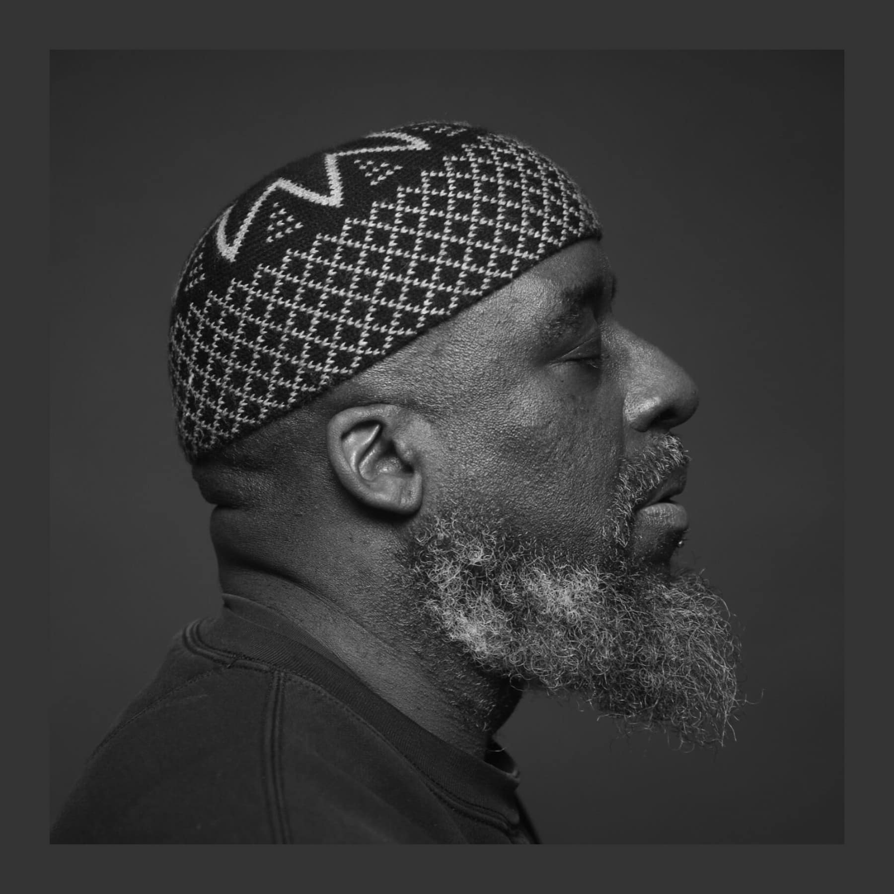 Black and white profile portrait of Ajamu X with eyes closed, in a patterned knitted cap, with a grey beard