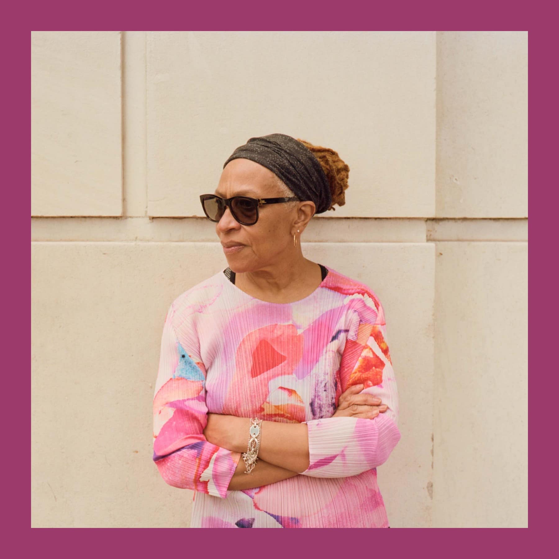 Veronica Ryan in sunglasses, a dark headwrap and a pink patterned top, arms folded against a pale stone wall