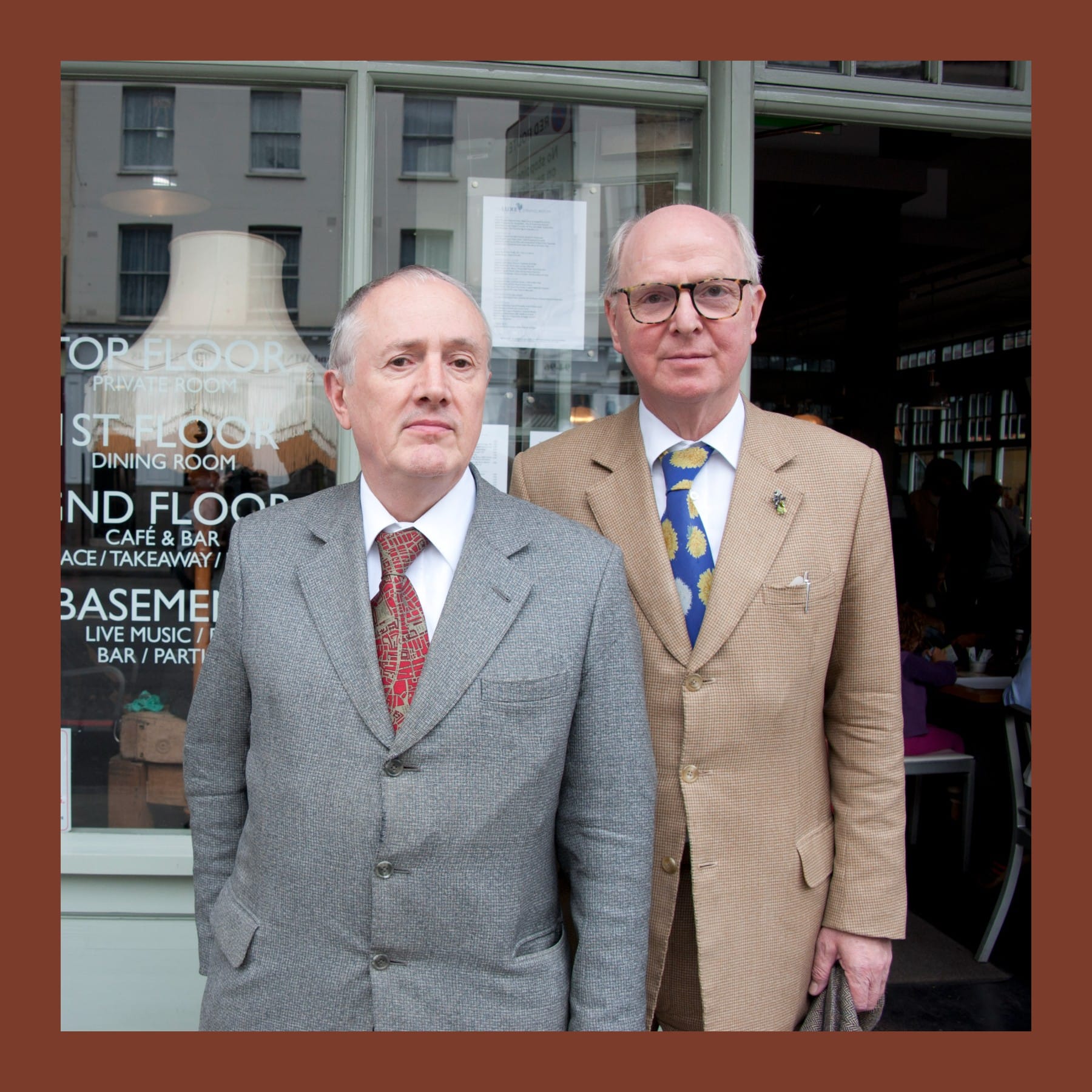 Gilbert and George in tweed suits standing side by side outside a café window in Spitalfields