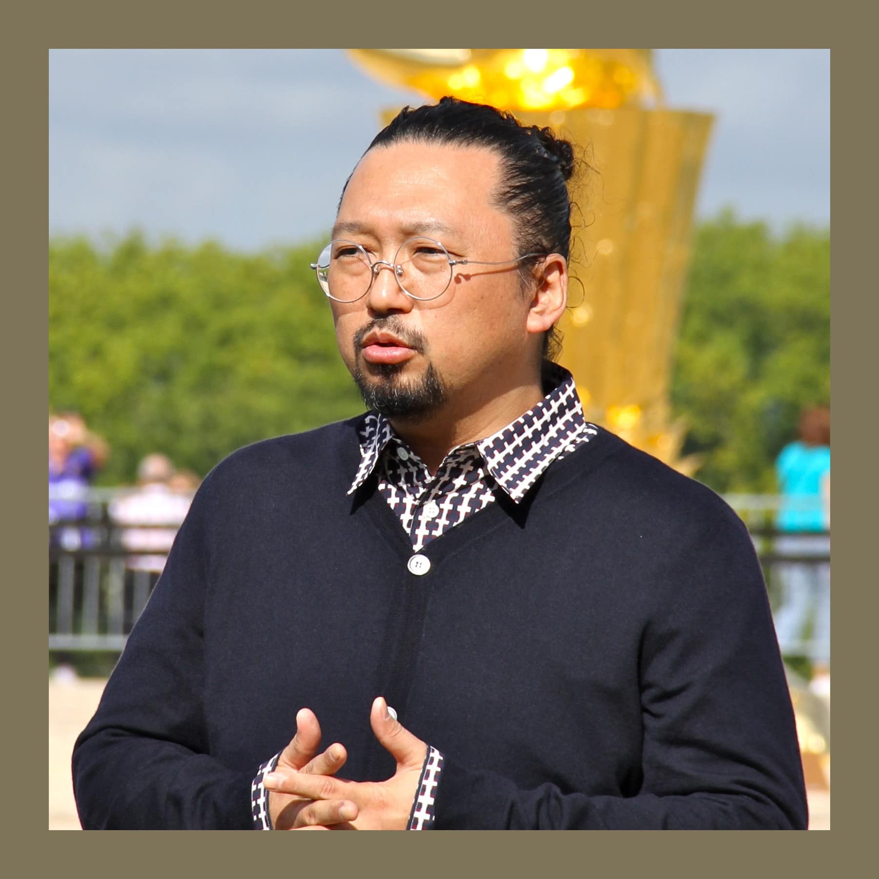 Takashi Murakami speaking outdoors at Versailles with a gold sculpture behind him