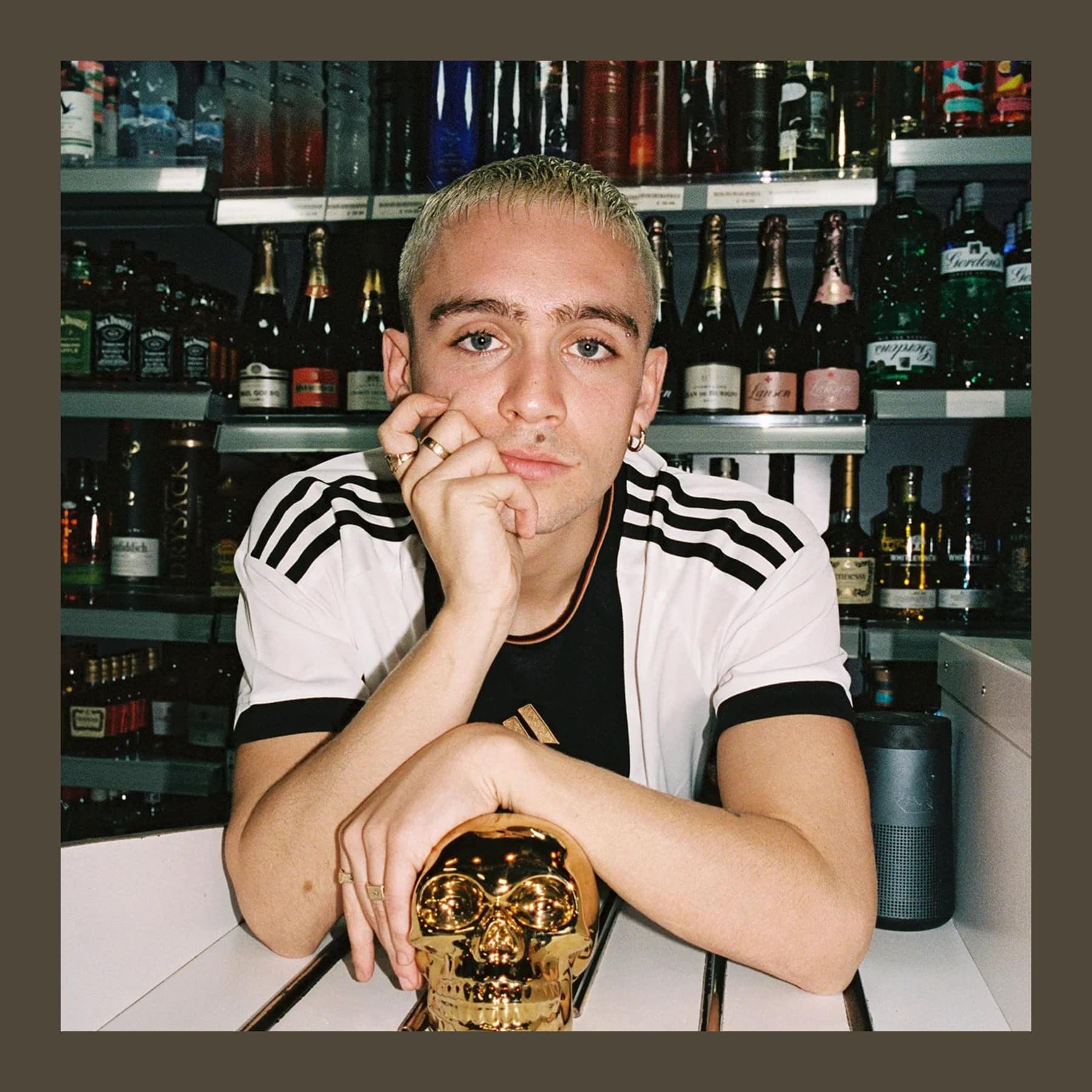 Corbin Shaw leaning on a counter in front of shelves of bottles, chin on his hand, beside a gold skull