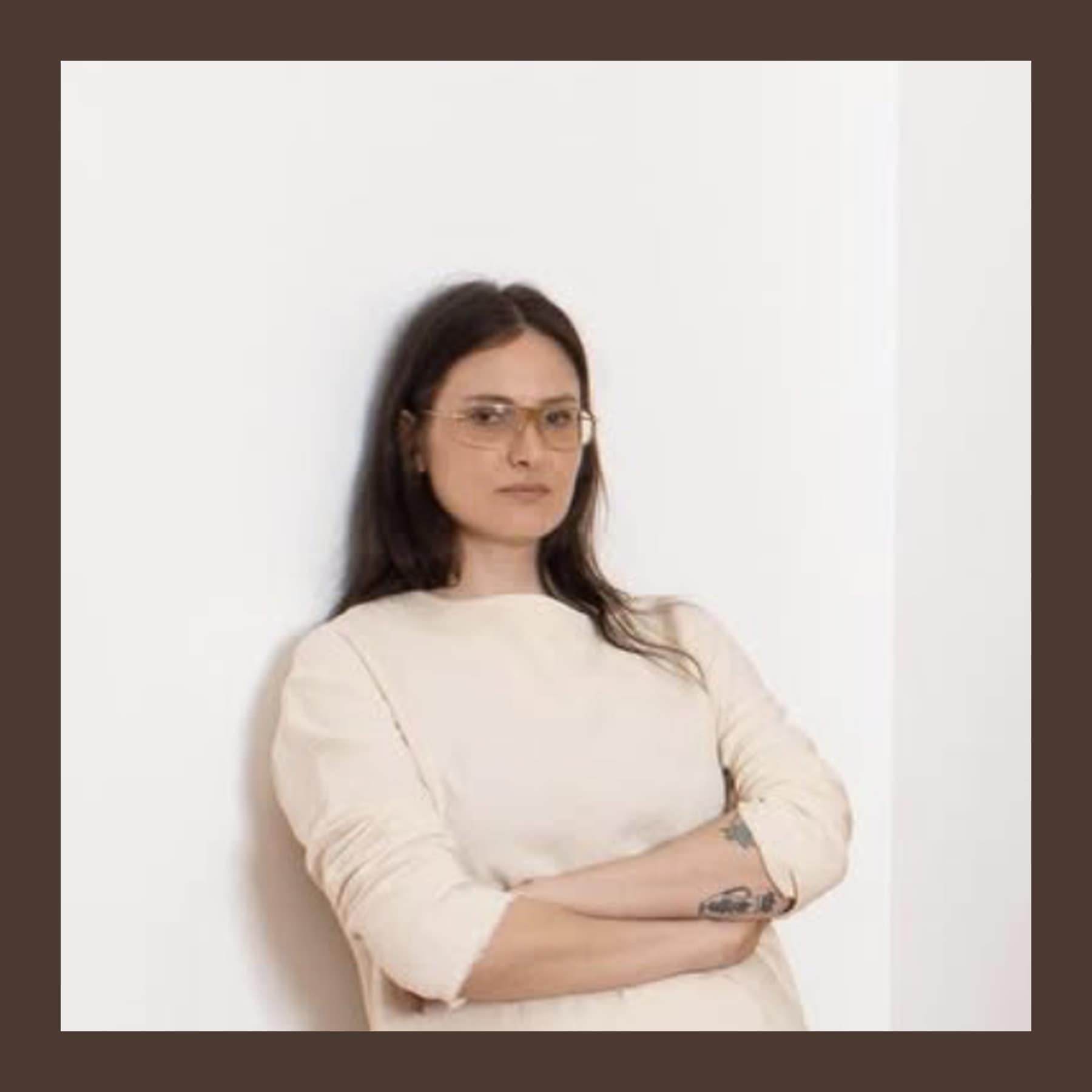 Olivia Drusin in tinted glasses and a cream top, leaning against a white wall with arms folded