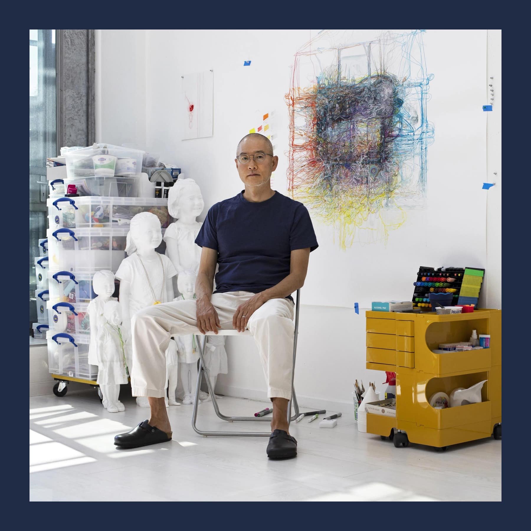 Do Ho Suh seated on a chair in his white studio, beside small white figures and a yellow storage unit