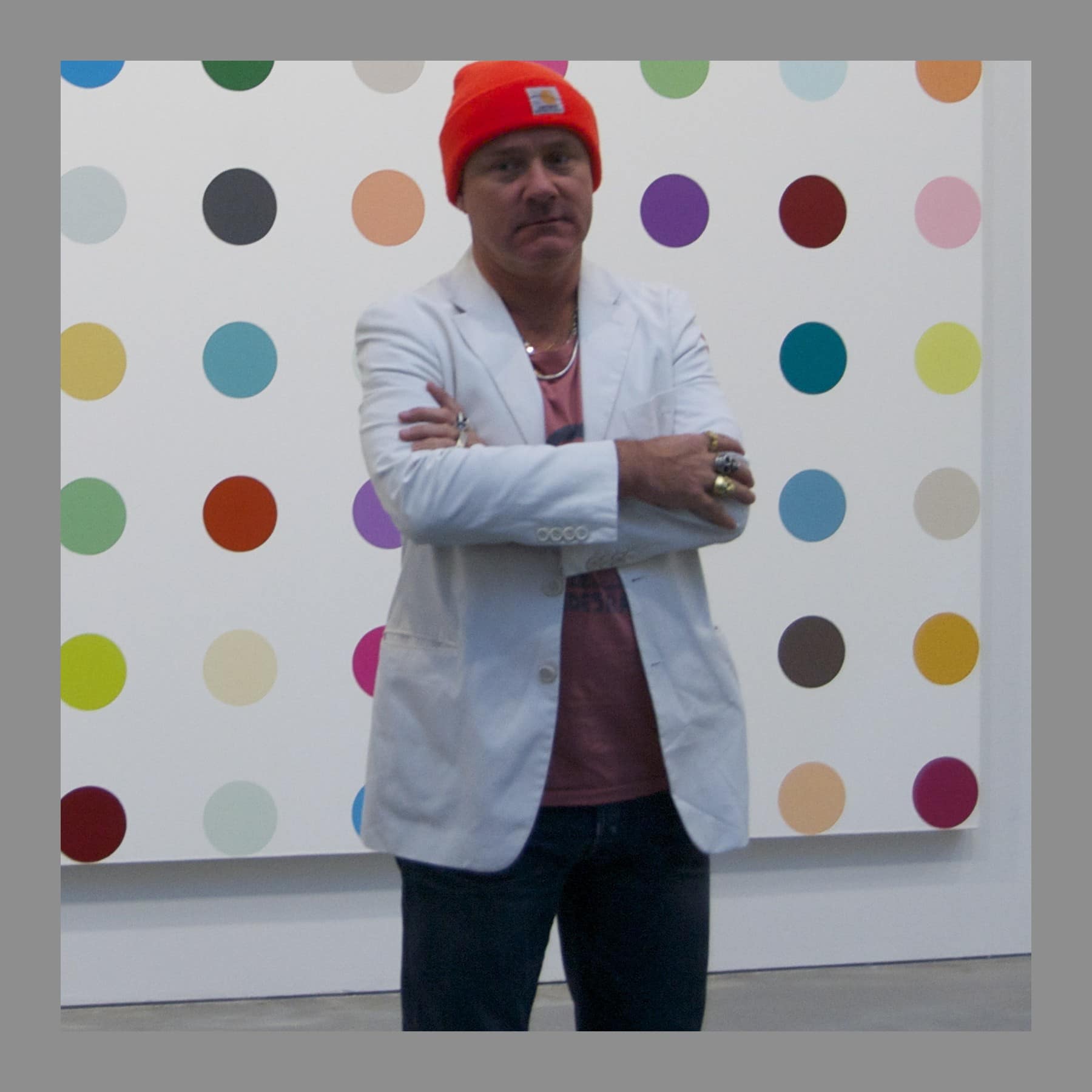 Damien Hirst in a red beanie, arms folded, in front of a spot painting