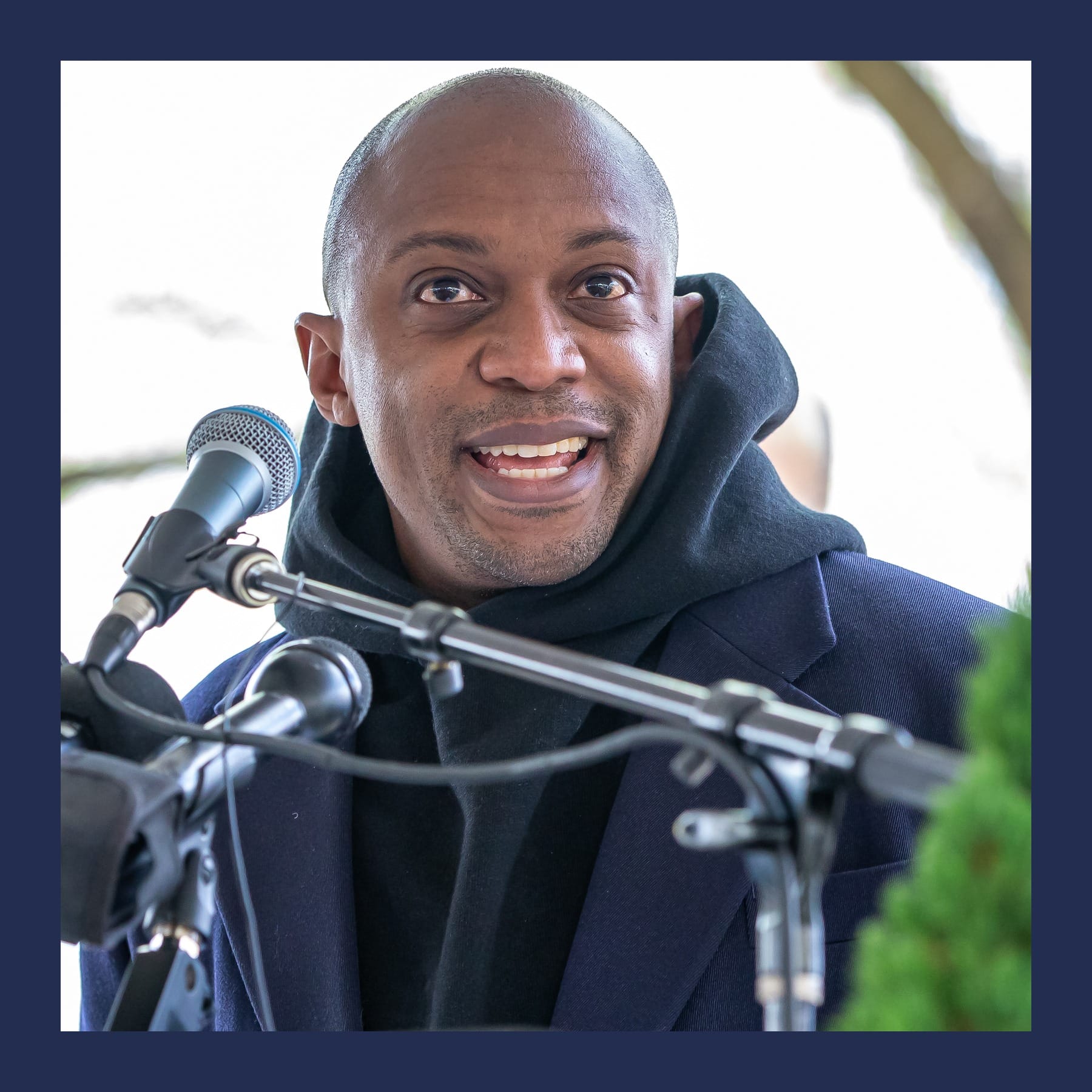 Hank Willis Thomas speaking at a microphone outdoors on Boston Common, in a dark coat and hoodie